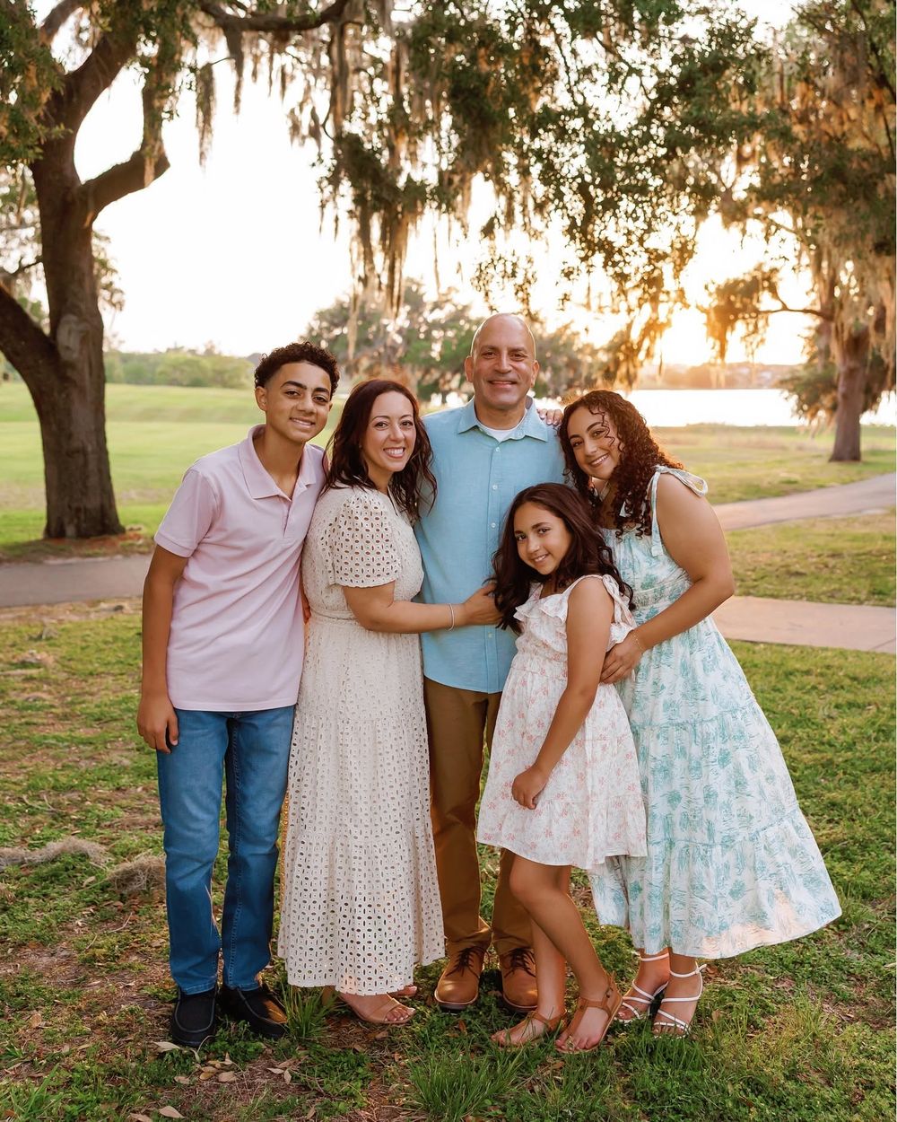 Family of five smiling outdoors under Spanish moss trees at golden hour in a park.