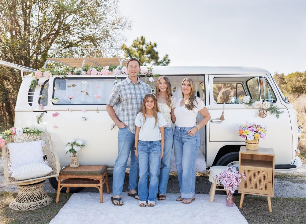 Family of four posing in front of a vintage white VW bus decorated with flowers, wearing matching jeans outfits.