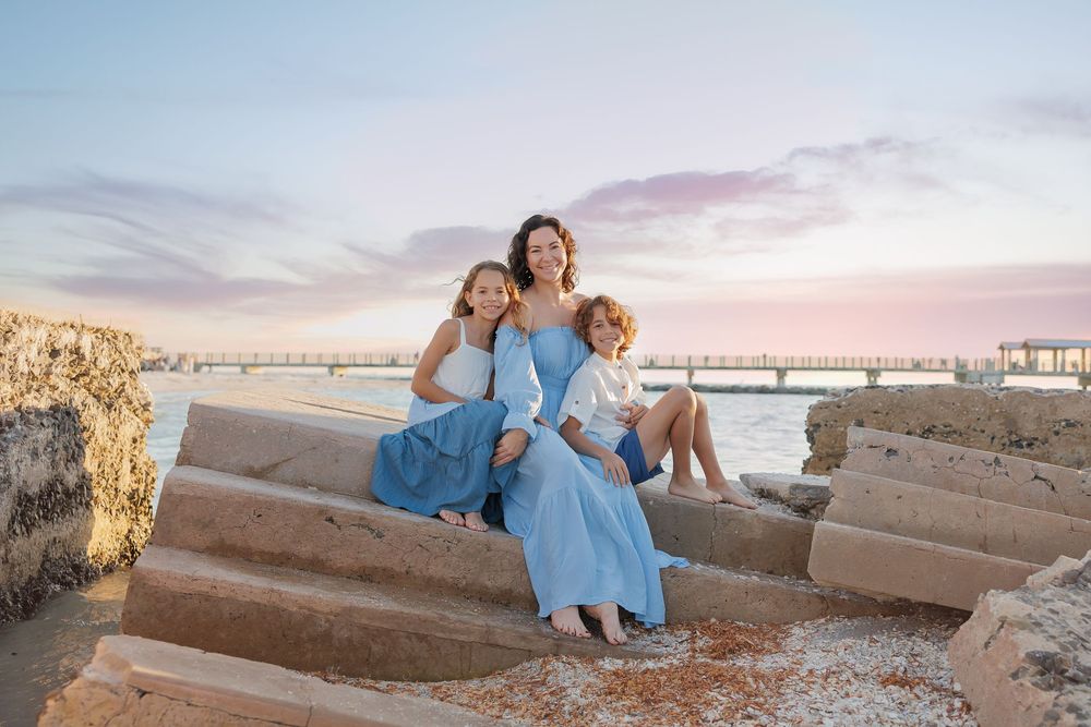 A mother and two children sit on stone steps by the waterfront at sunset, all wearing blue outfits.