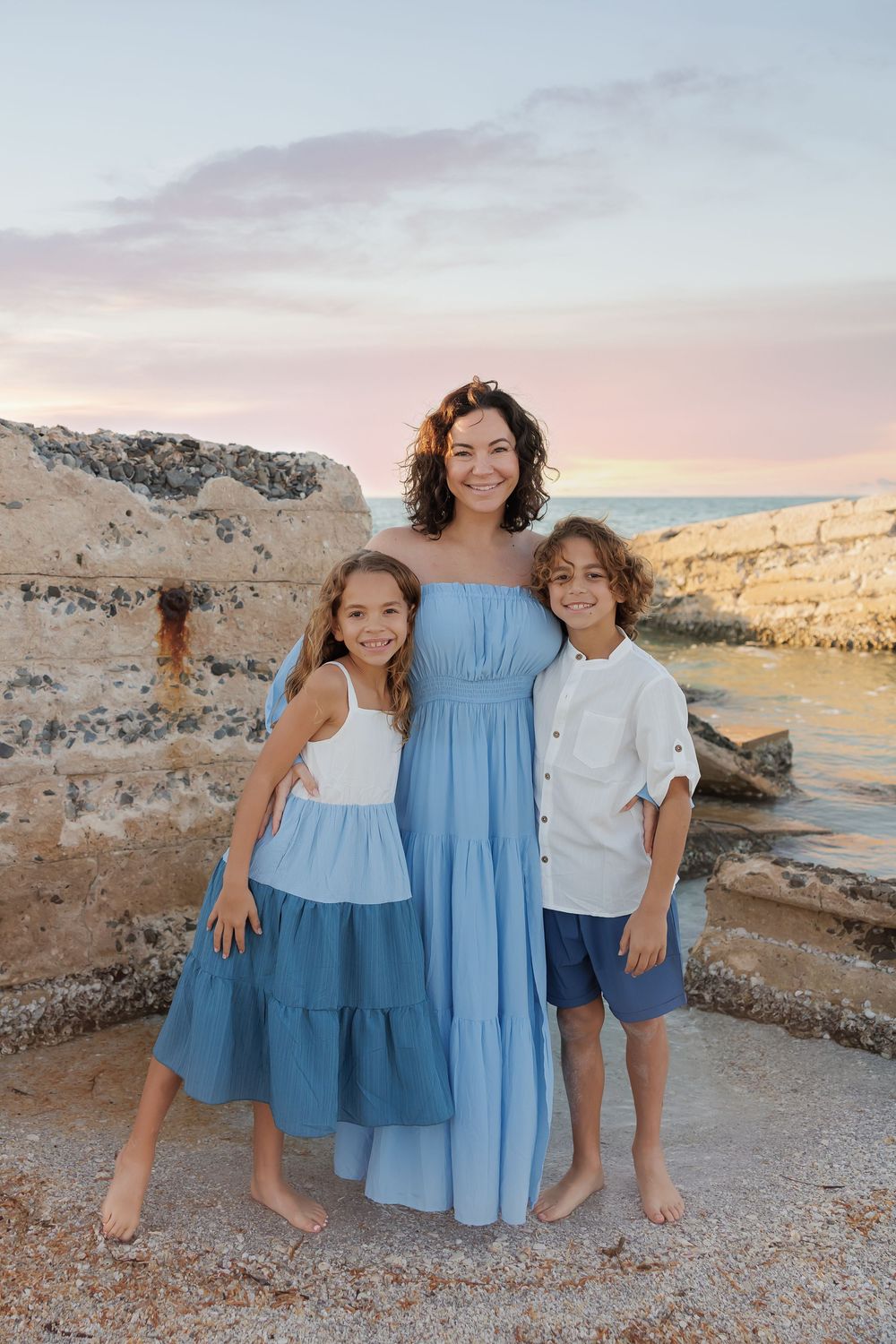 A woman in a blue dress poses with two children by rocky seaside ruins at sunset.