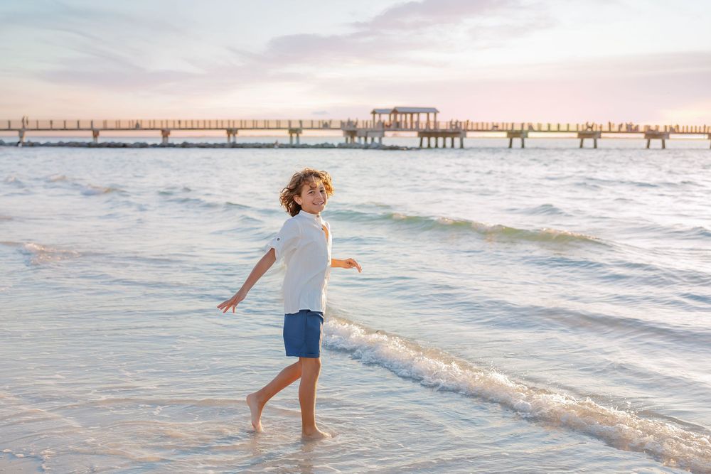 A young boy with curly hair walks along the ocean shoreline at sunset, wearing a white shirt and blue shorts.