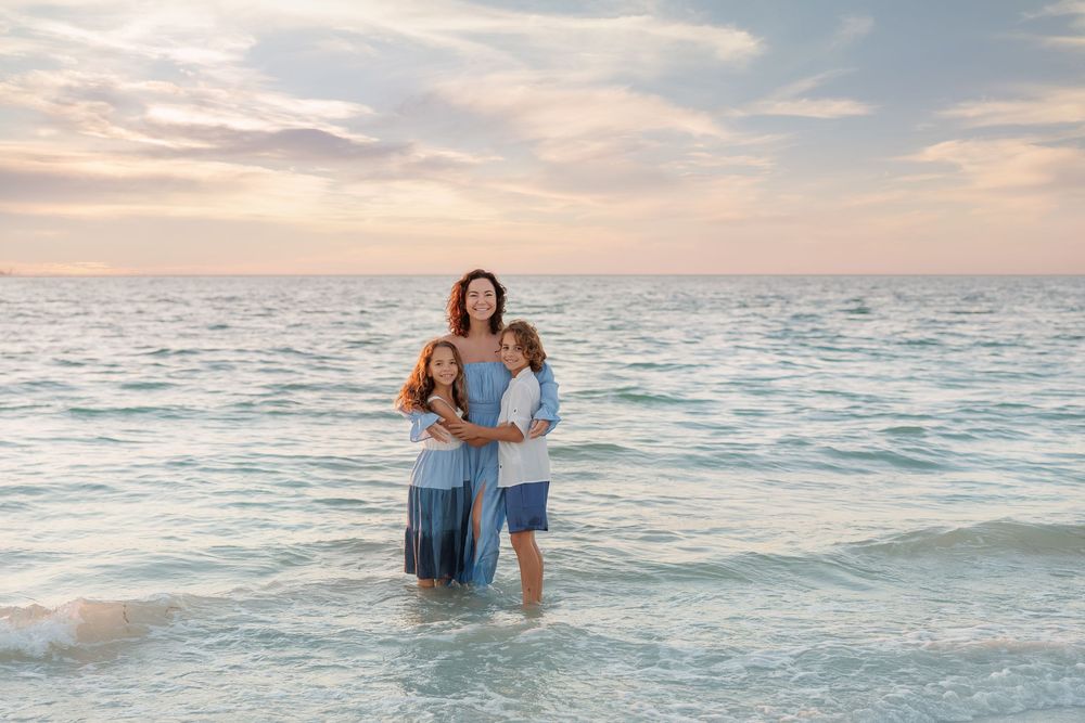 Woman and two children standing in ocean waves at sunset, wearing blue outfits, smiling at camera.