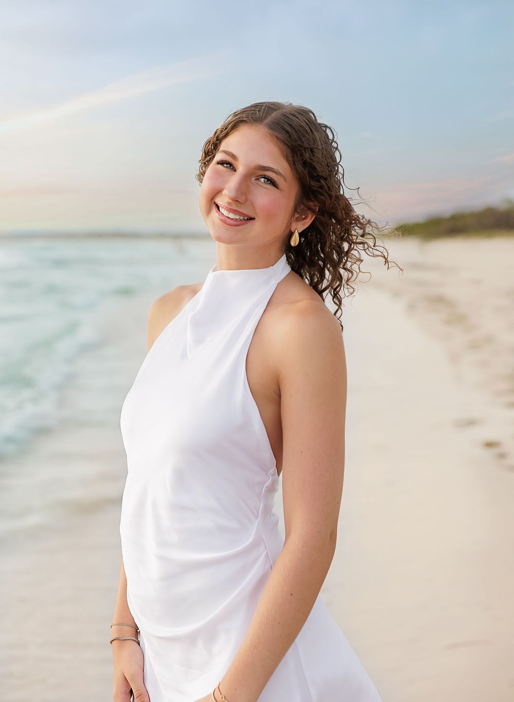 Young woman in white halter dress smiling on a white sand beach at sunset.