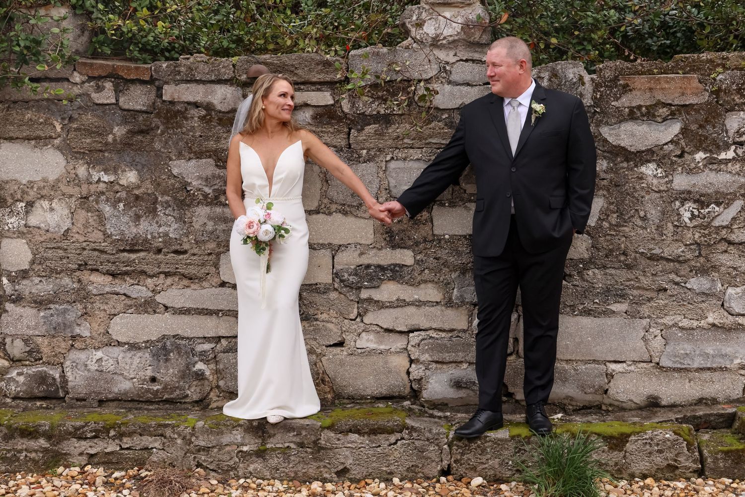 Bride and groom holding hands smiling in front of a rustic stone wall, bride holding a white bouquet.