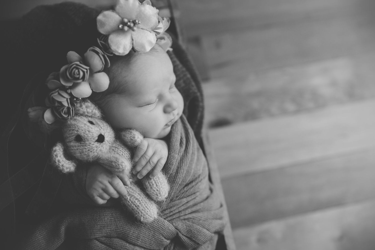 Black and white newborn photo of sleeping baby with floral headband cuddling a knit teddy bear.