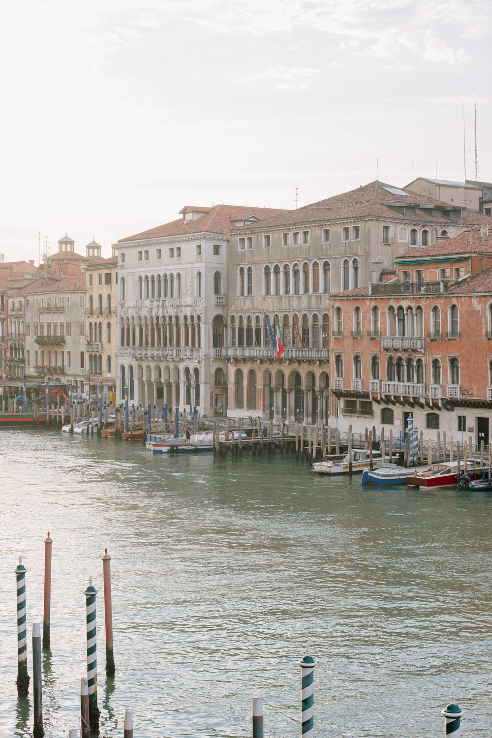 Grand Canal Venice with historic Renaissance palazzos, moored boats, and wooden dock posts reflected in calm waters.