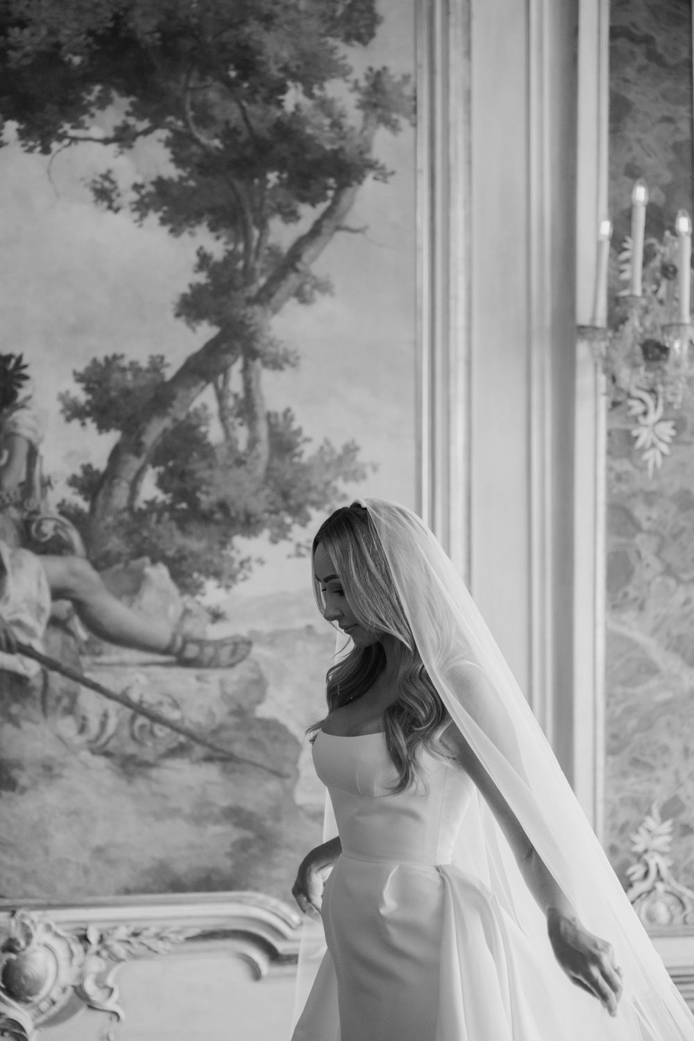 Black and white photo of a bride in a strapless gown and veil standing by a window with ornate mural backdrop.