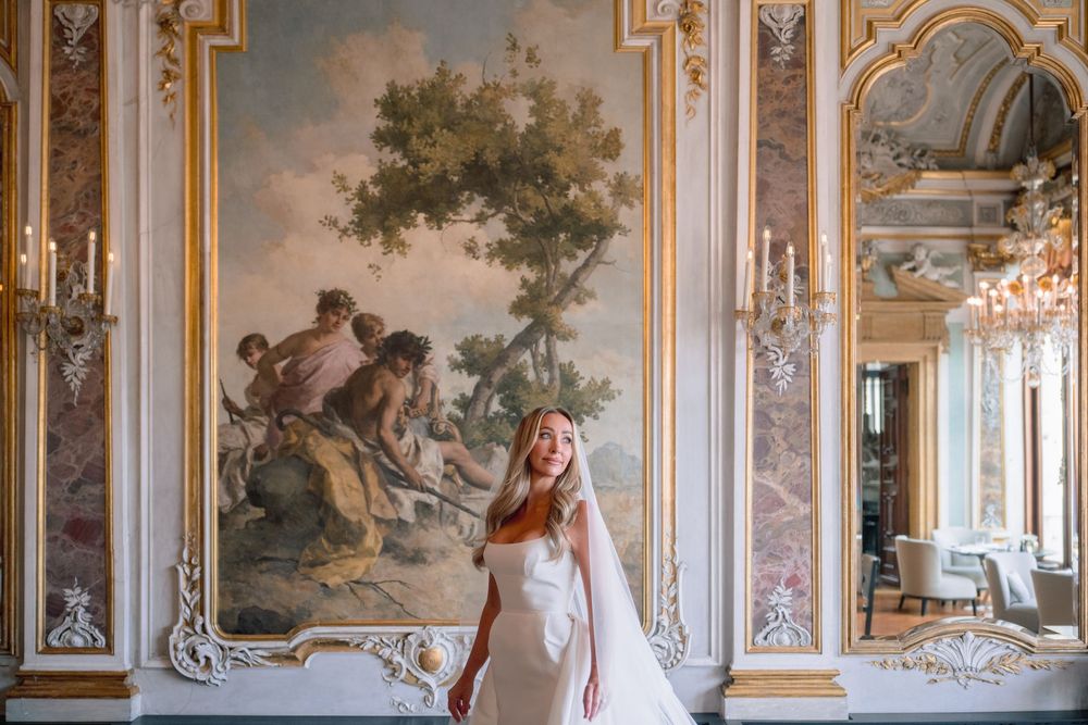 Bride in white gown poses before ornate gold-framed mural in elegant baroque ballroom with crystal chandeliers.