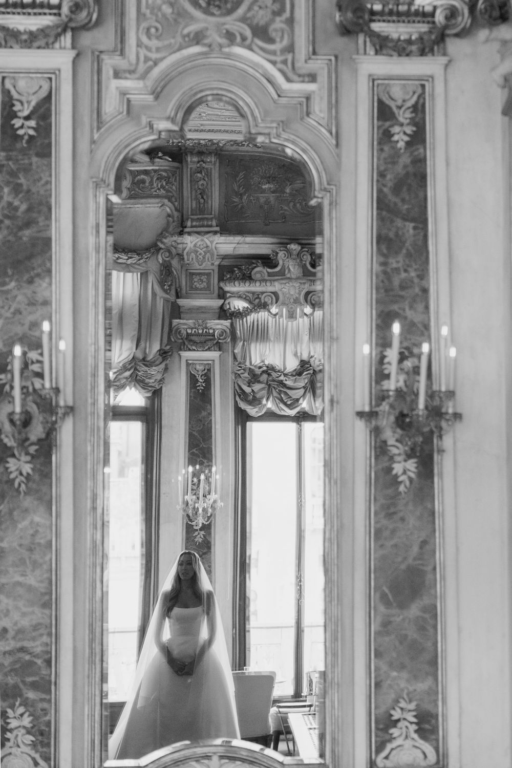 Black and white photo of a bride in wedding dress reflected in ornate baroque mirror in elegant ballroom.