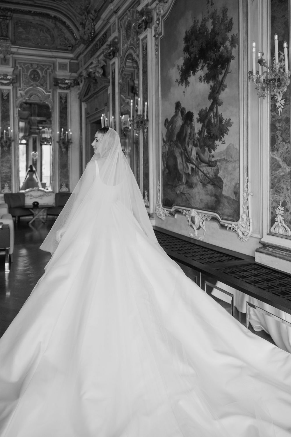 Bride in elegant white gown with long train and veil stands in ornate baroque ballroom with murals and candelabras.