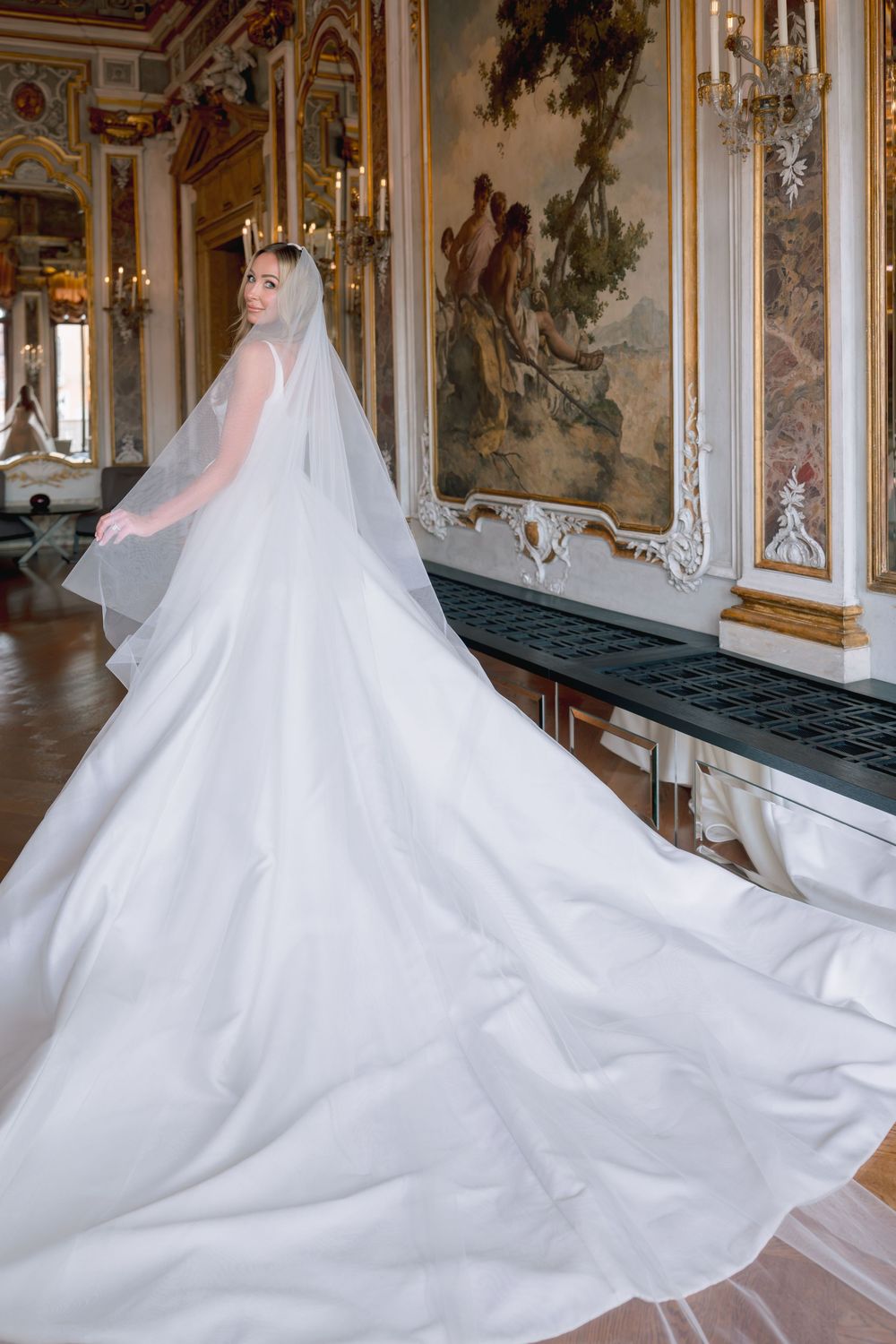 Bride in elegant white gown with long train and veil poses in ornate gilded hall with classical paintings.