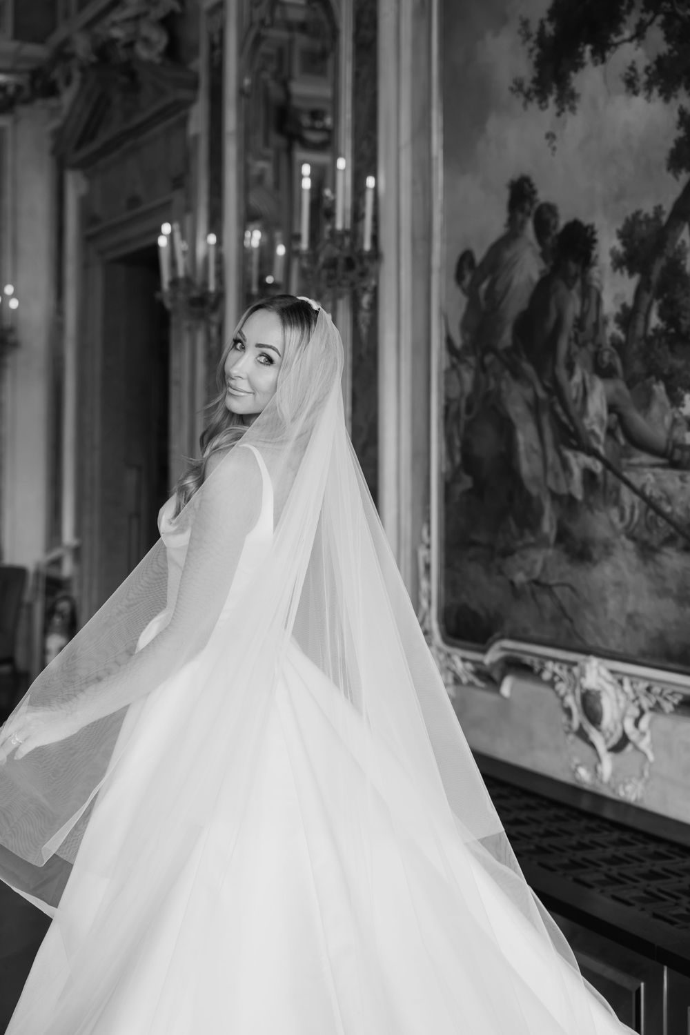 Black and white photo of a smiling bride in a flowing gown and veil in an ornate ballroom.
