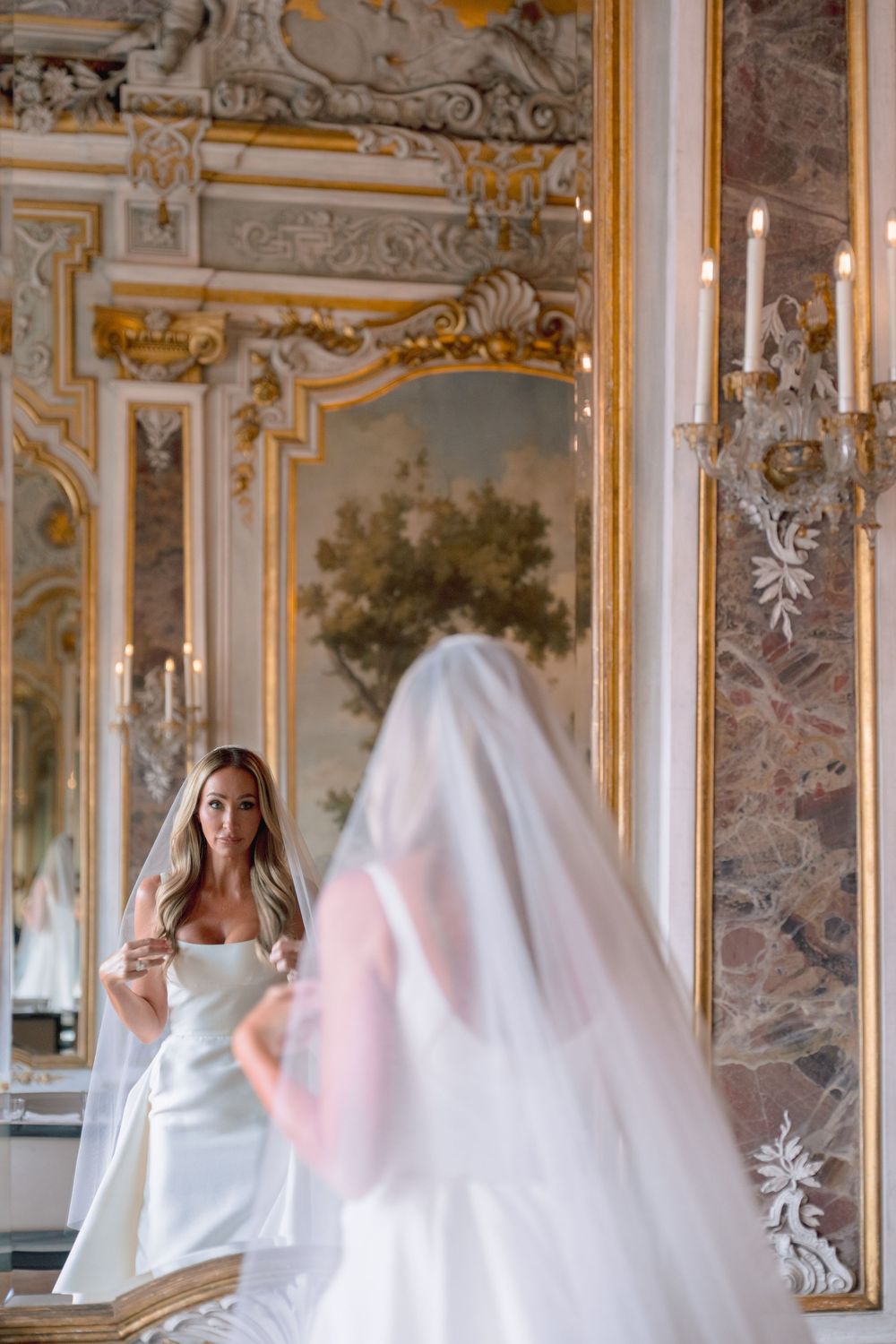 Bride in white gown and veil admires herself in ornate gold-framed mirror in elegant baroque palace.