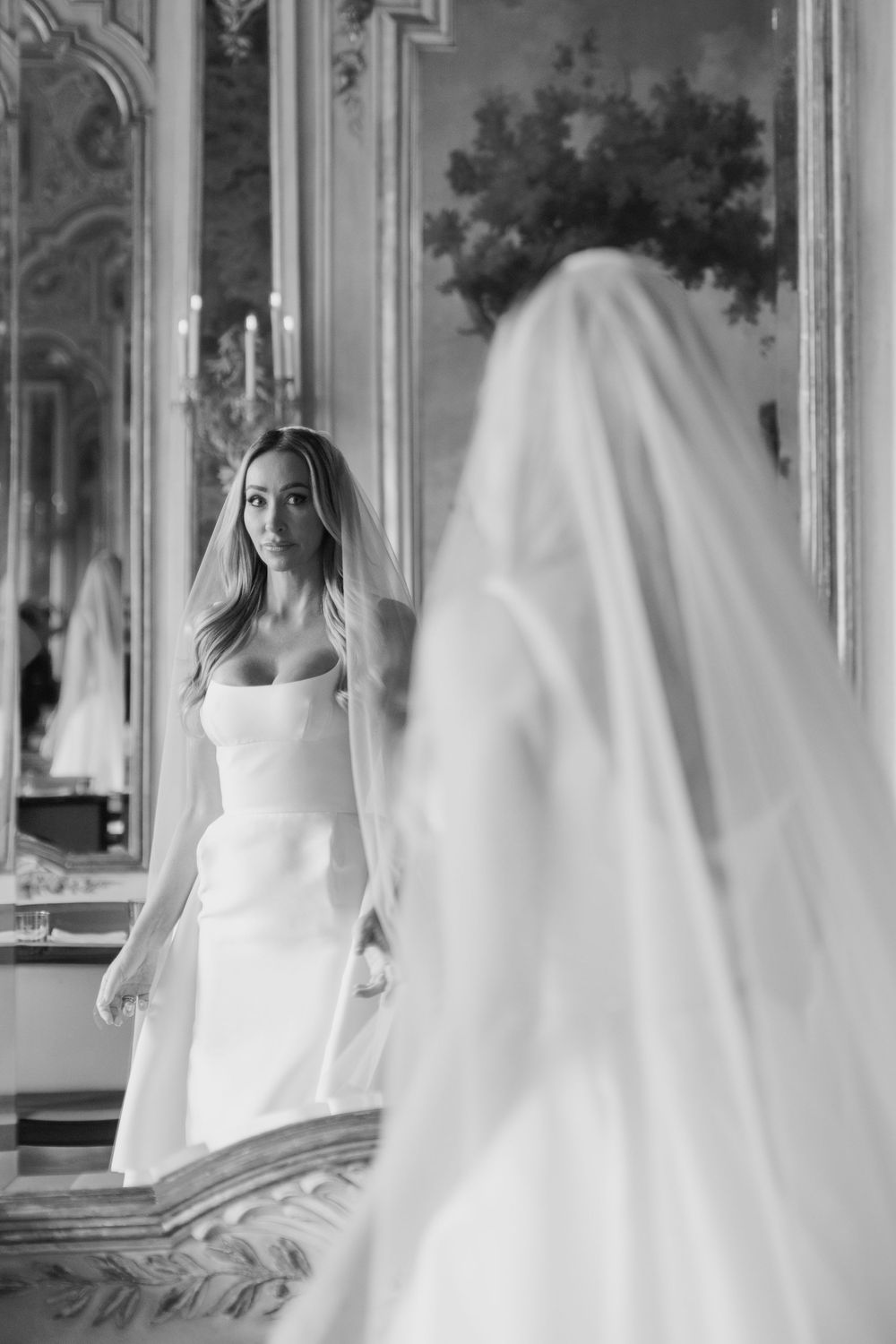 Bride in elegant strapless white gown and veil gazes at her reflection in an ornate gold mirror.