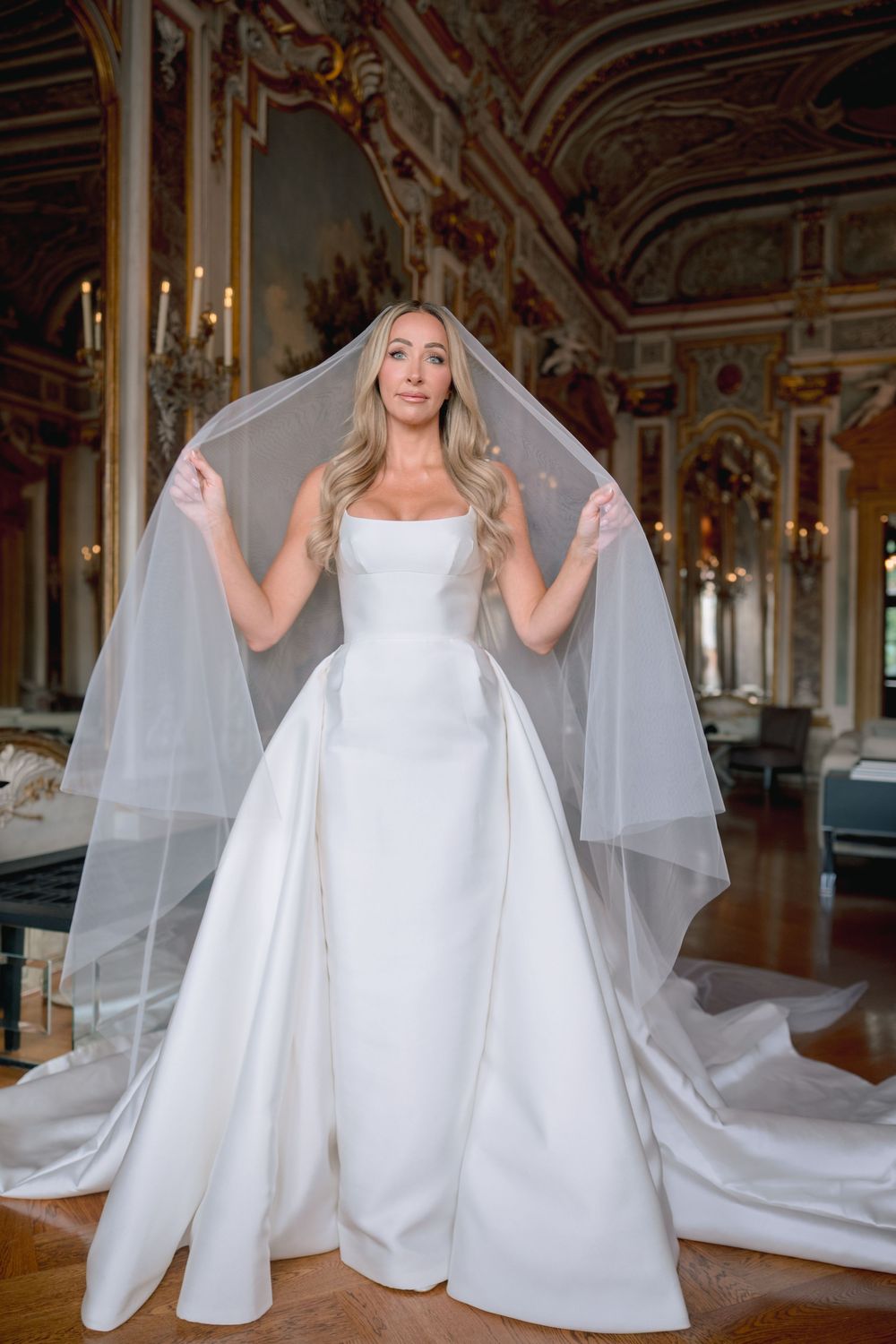Bride in elegant white satin gown with flowing veil in ornate gilded ballroom.