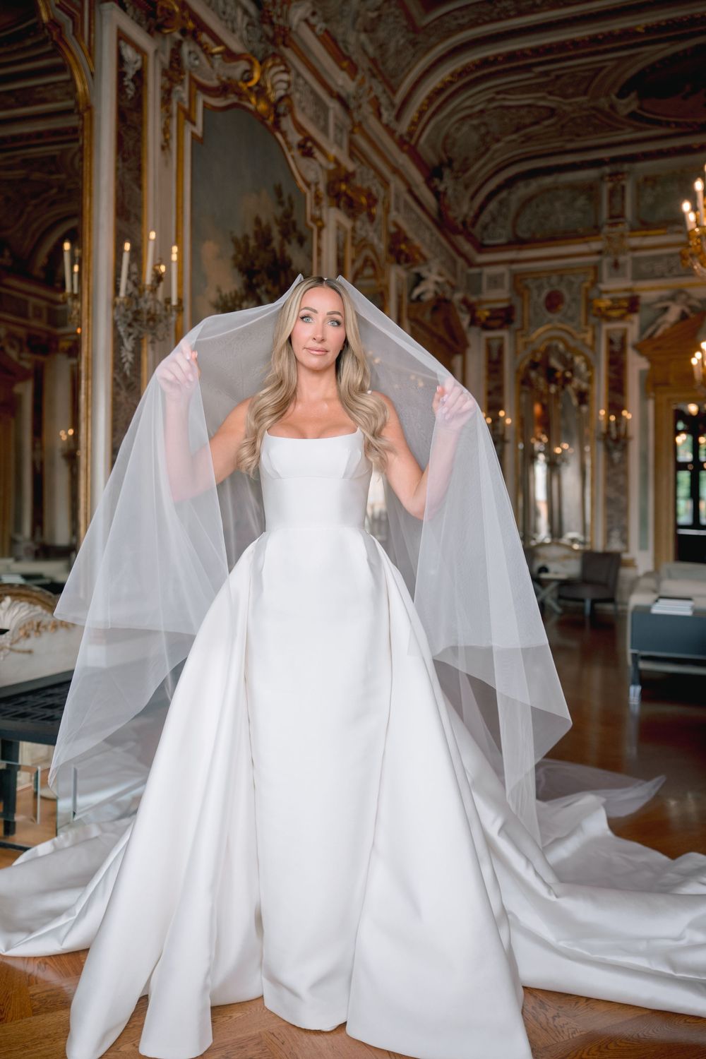 Bride in a strapless white satin ball gown holding a flowing tulle veil in an ornate gilded ballroom.