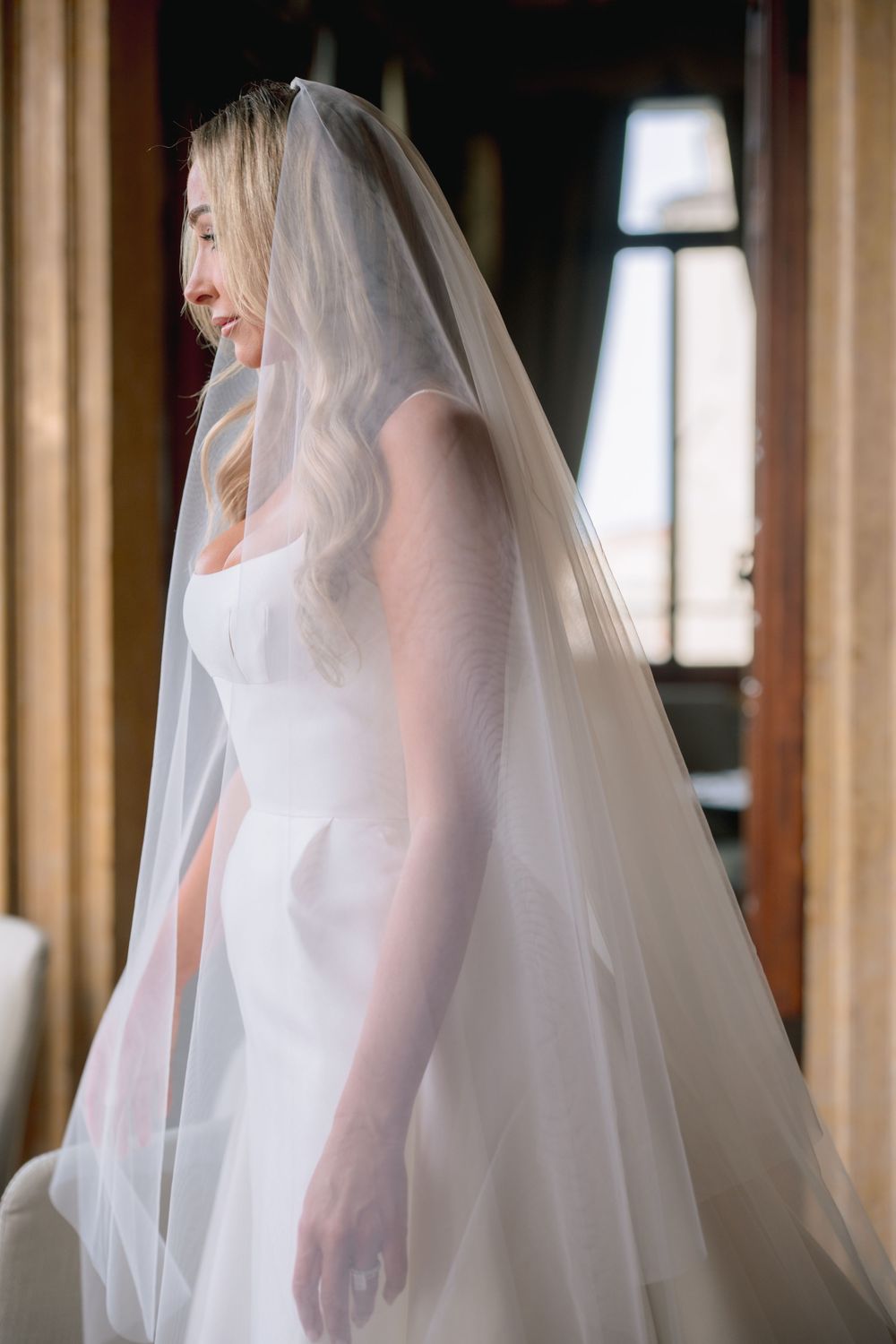 Bride in white wedding dress and long flowing veil standing by window in elegant venue.