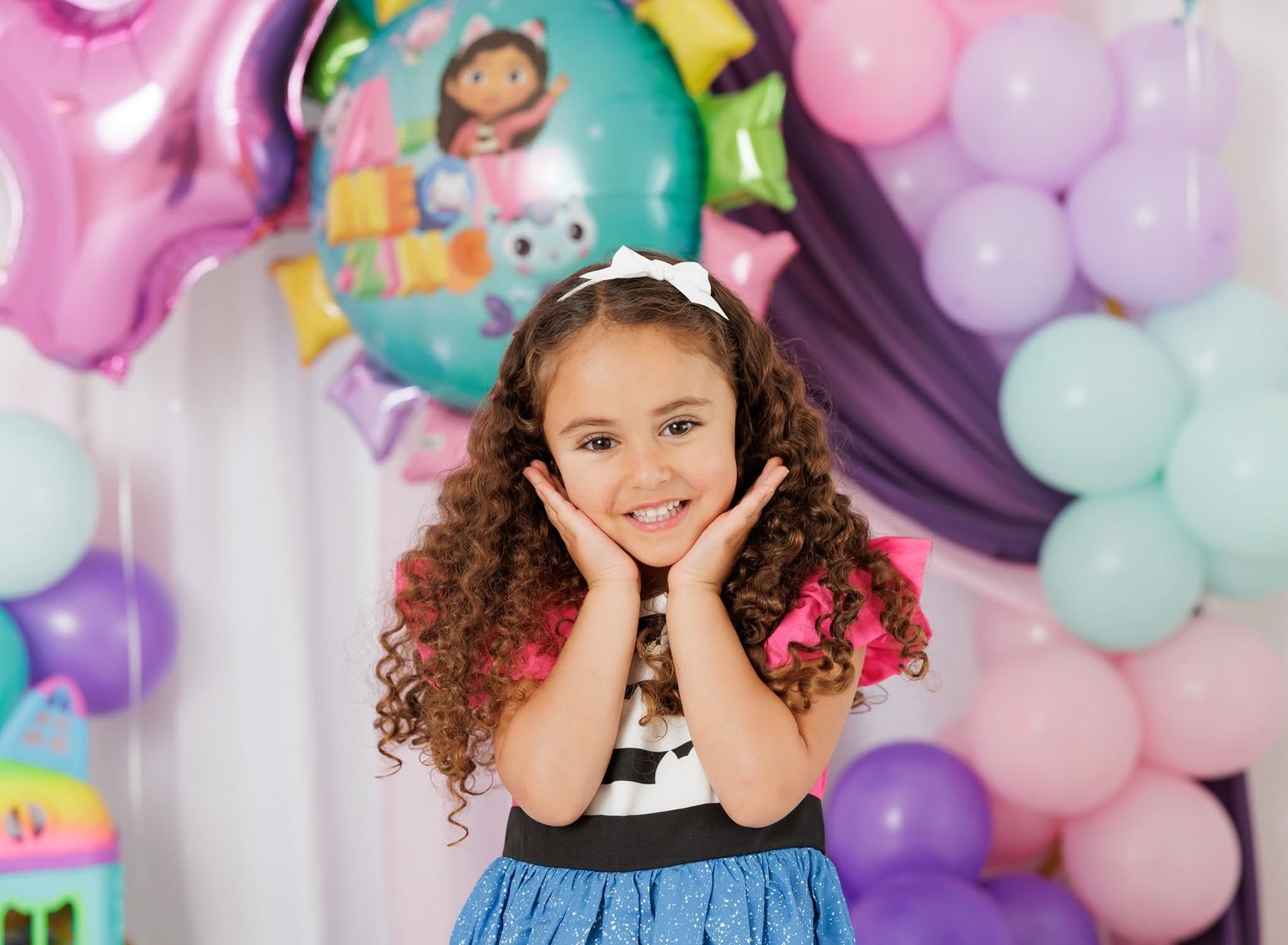 A smiling toddler girl in a tan ruffled dress sits on a white floor with a balloon in a bright studio setting.