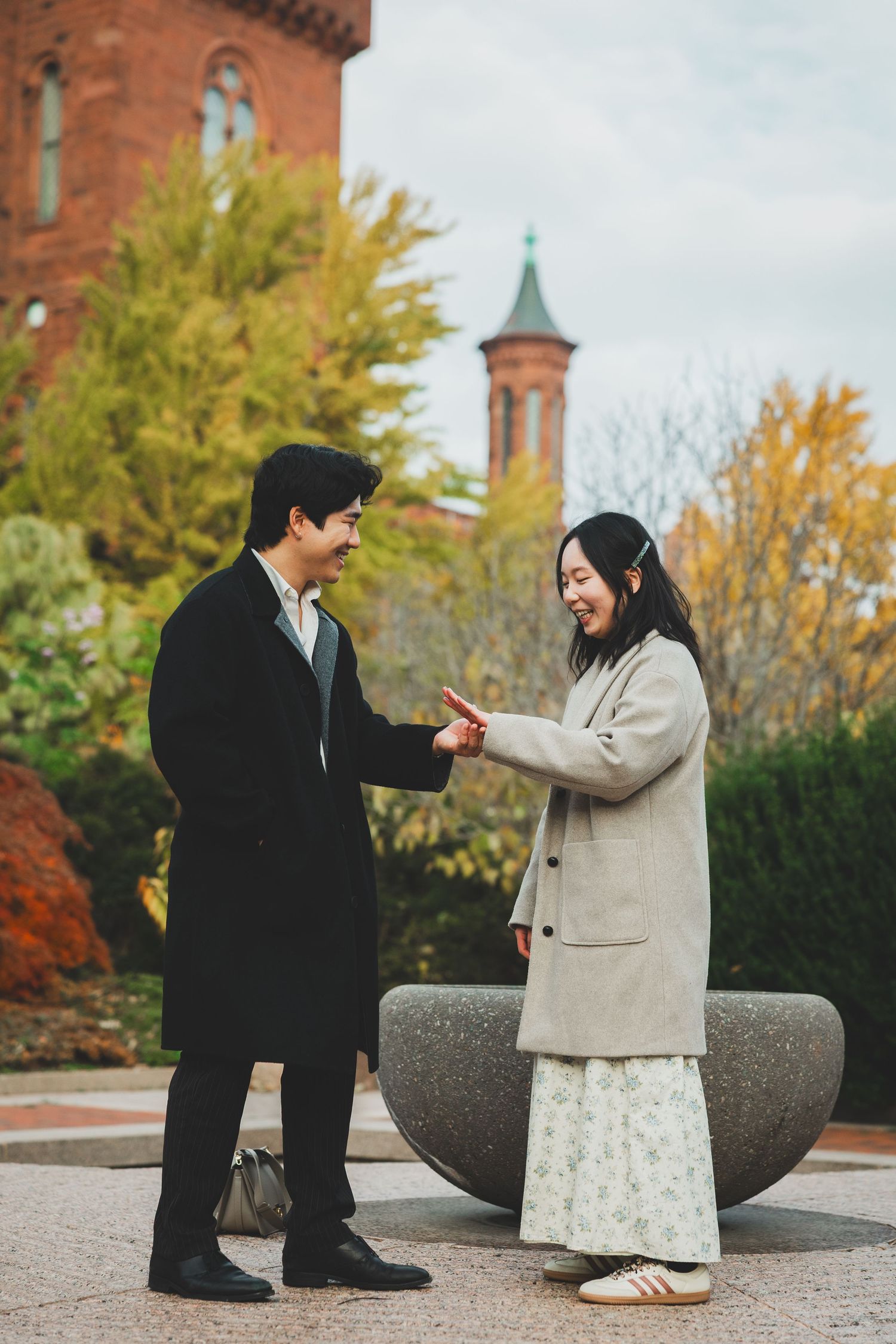 Couple holding hands and smiling outdoors near a red brick building with a tower, surrounded by autumn foliage.
