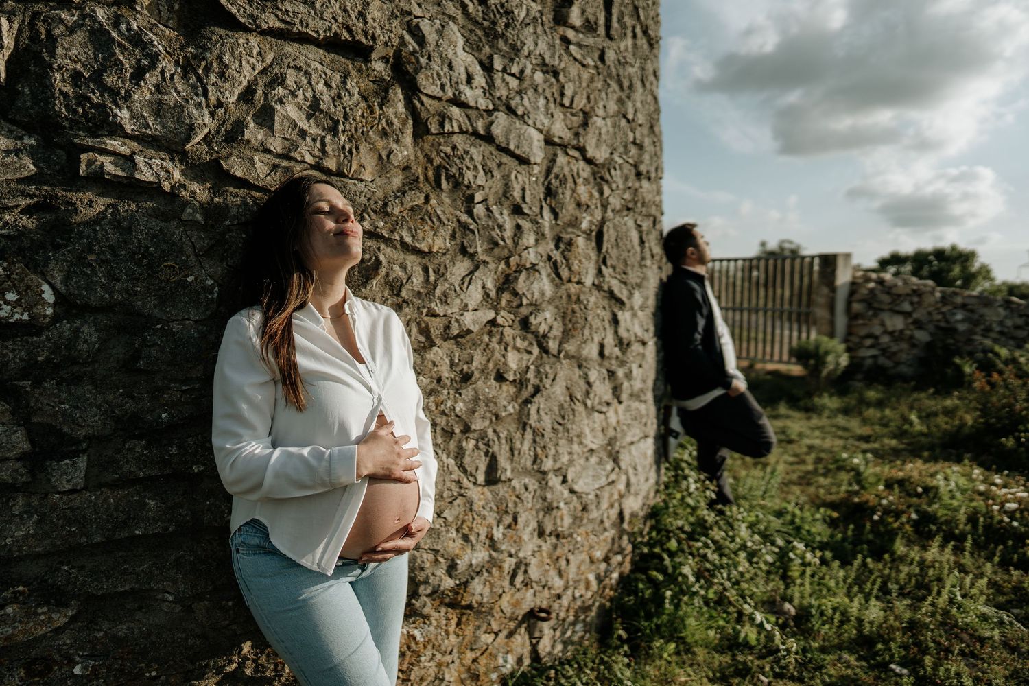 A person in a white blouse poses against a stone wall in a natural outdoor setting with dramatic cloudy skies.