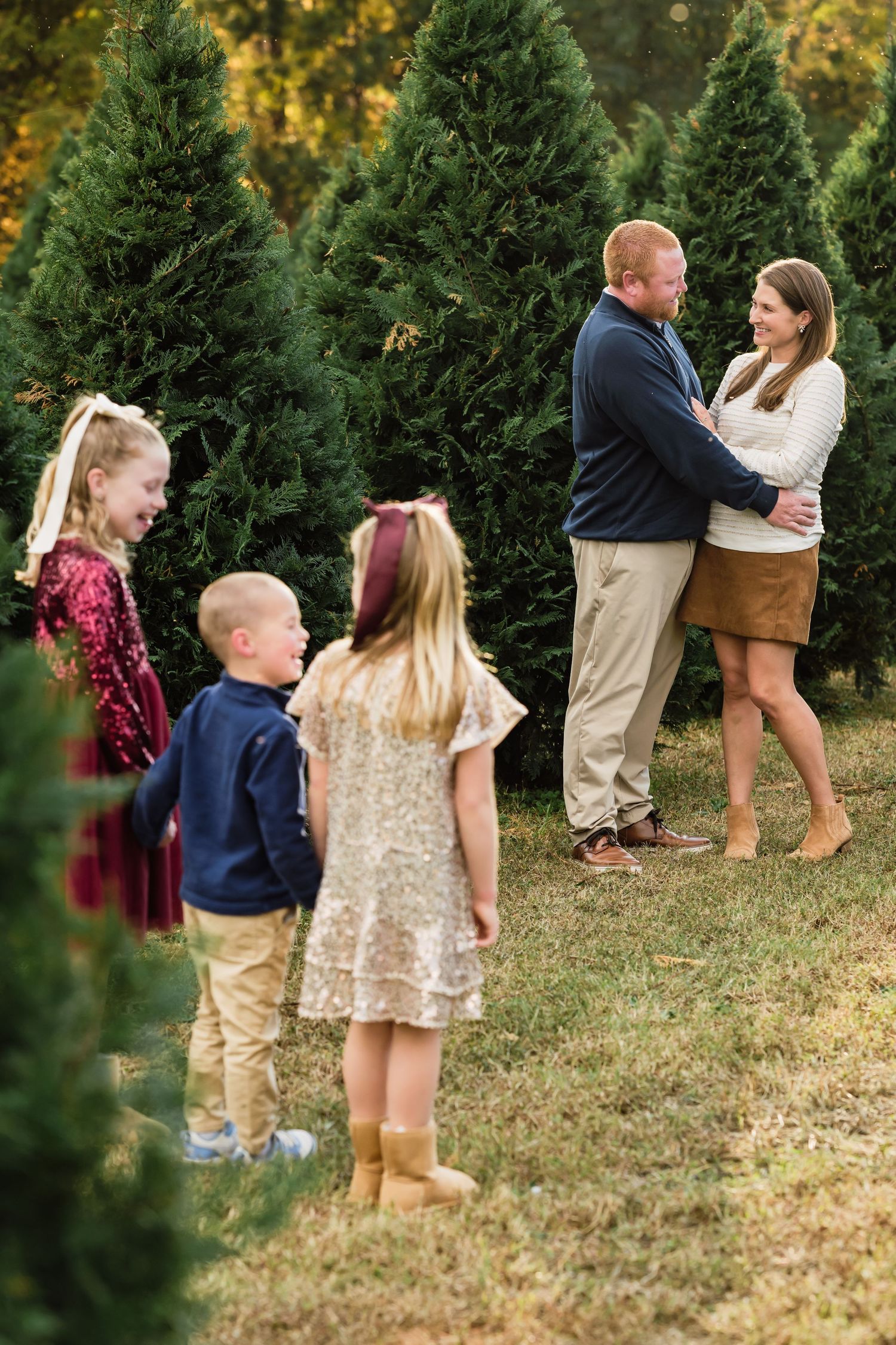 Family holiday photo session at a Christmas tree farm with couple embracing and three children playing nearby.