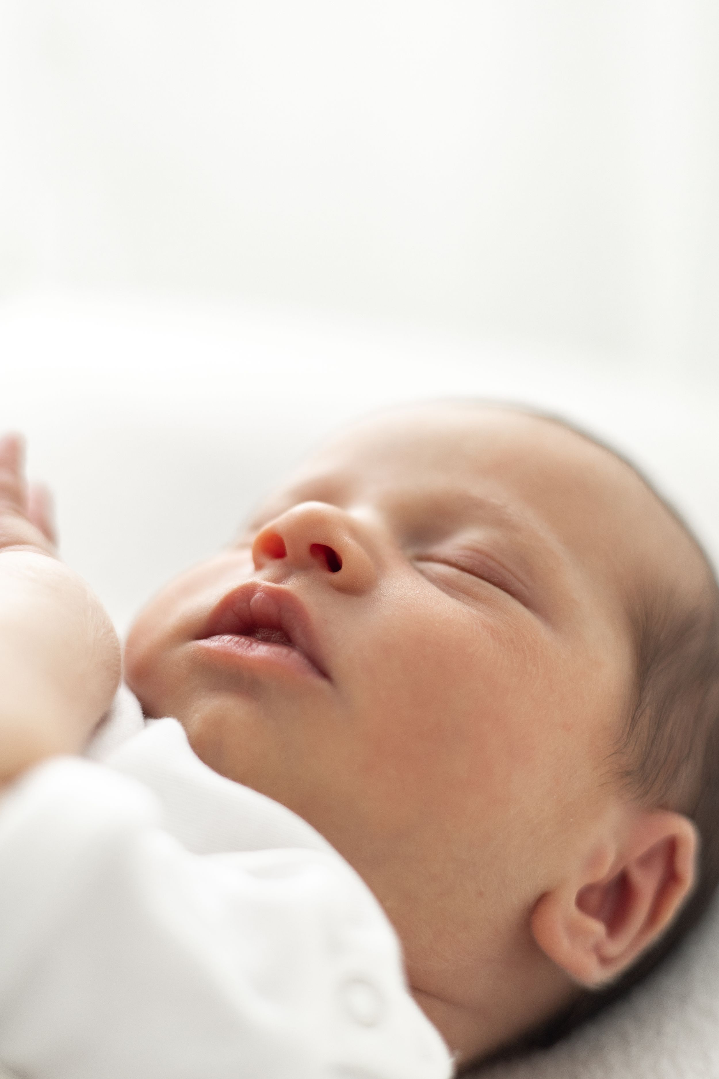 Peaceful newborn baby sleeping on a white surface, wearing a white onesie, and tiny hands visible Newborn photographer Nottingham