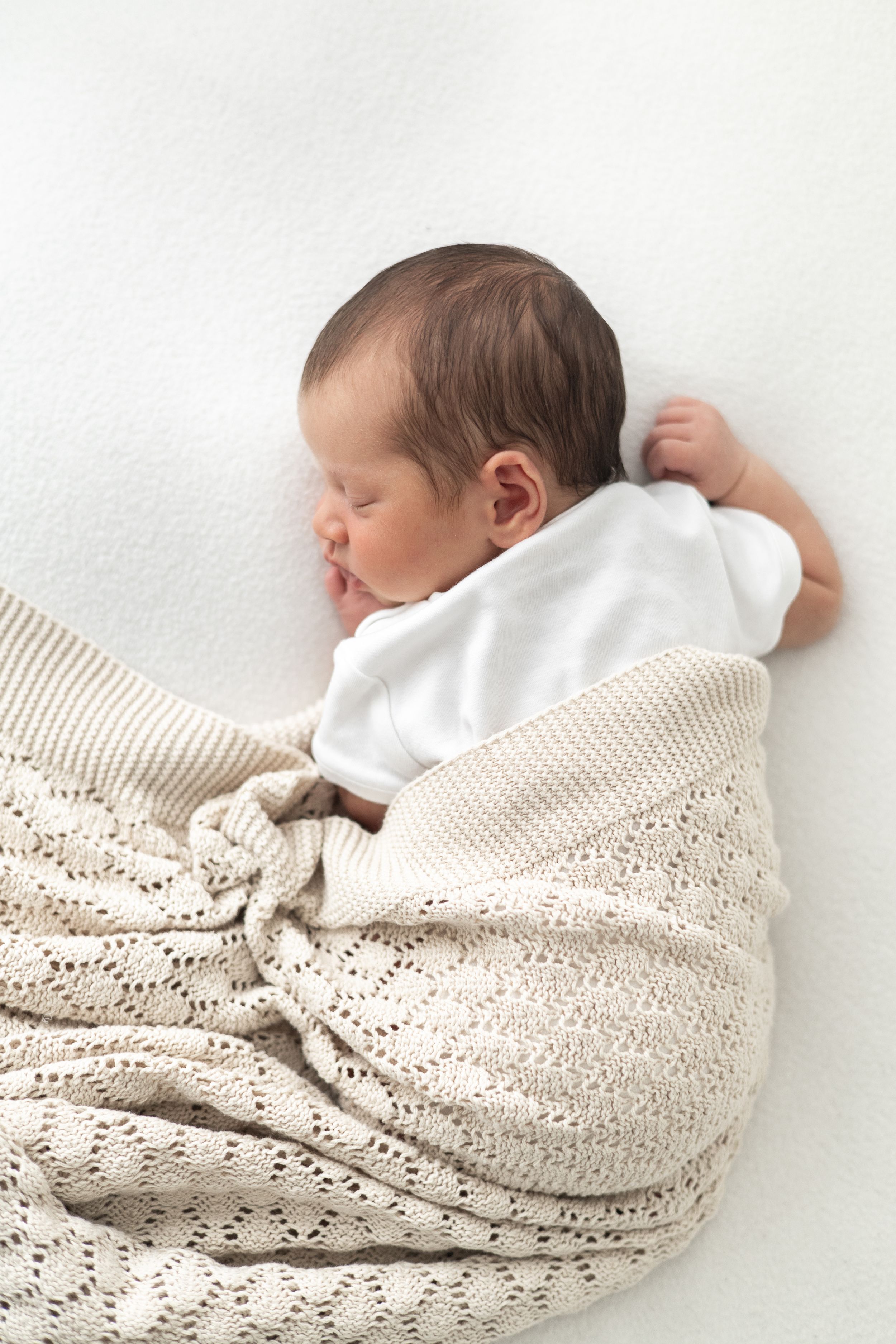 Newborn baby sleeping peacefully wrapped in a soft cream blanket wearing white onesie on a white background Newborn photographer Nottingham