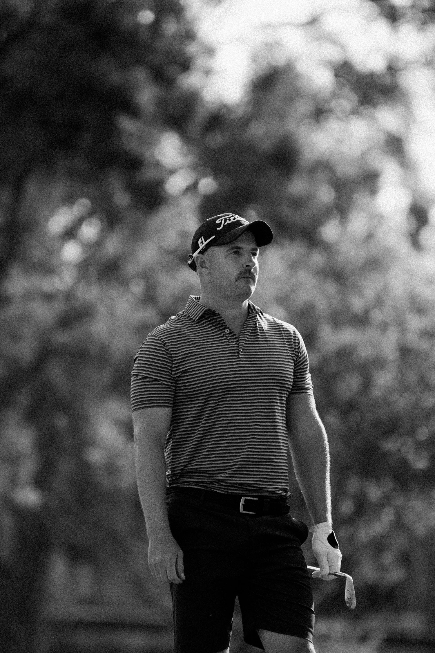 Black and white photo of a male golfer wearing a Titleist cap and striped polo shirt on a golf course.