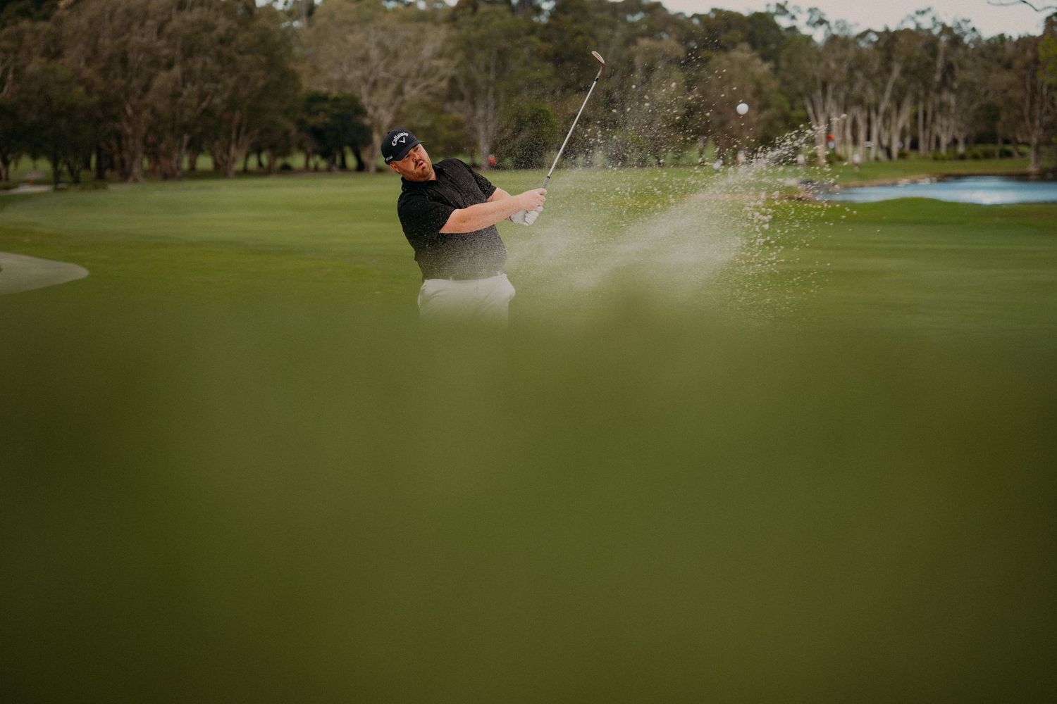 Golfer hits bunker shot sending sand flying on lush green golf course.