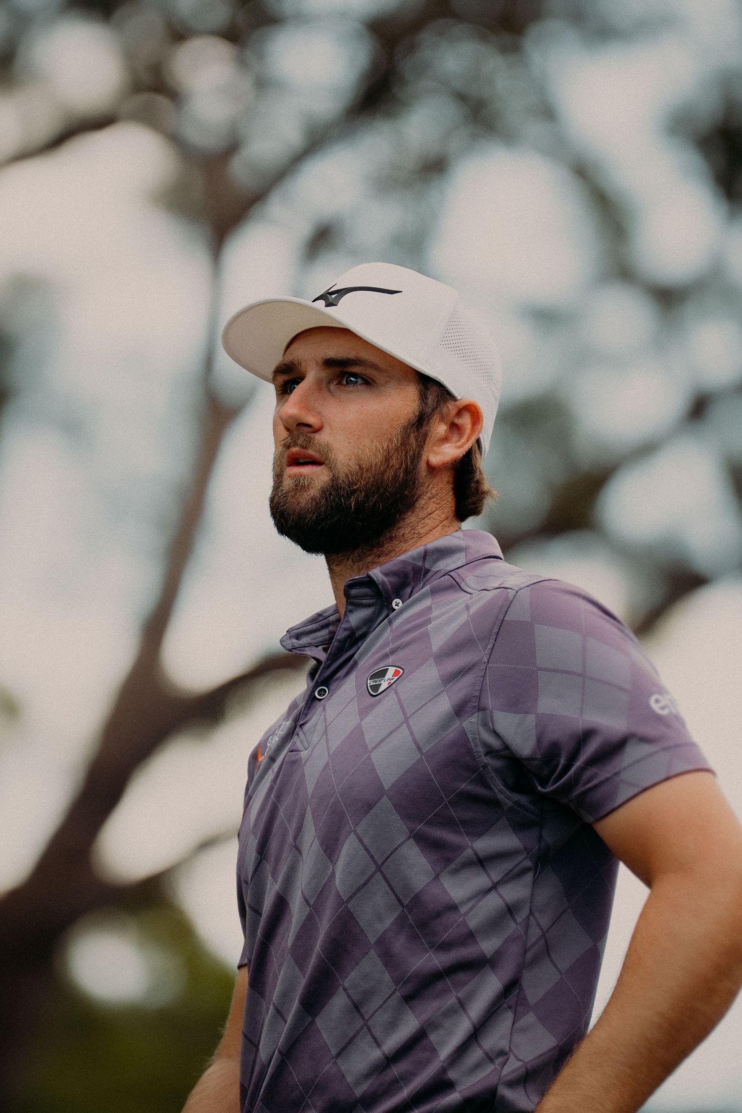 Young man with beard wearing purple argyle polo shirt and white golf cap standing outdoors on golf course.