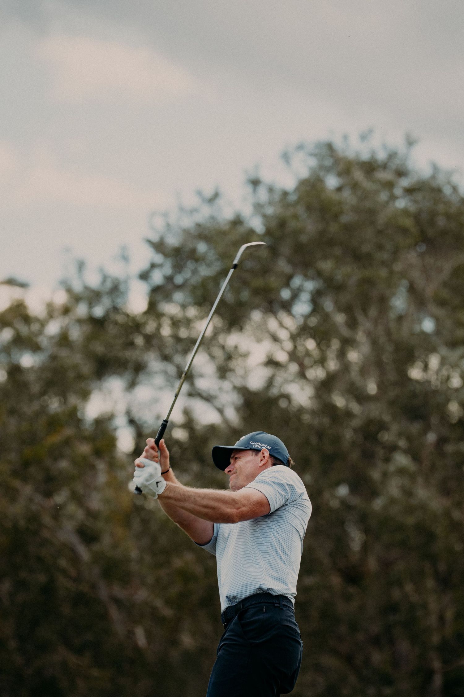 Golfer in white shirt and cap follows through on a powerful swing on a golf course with trees in background.