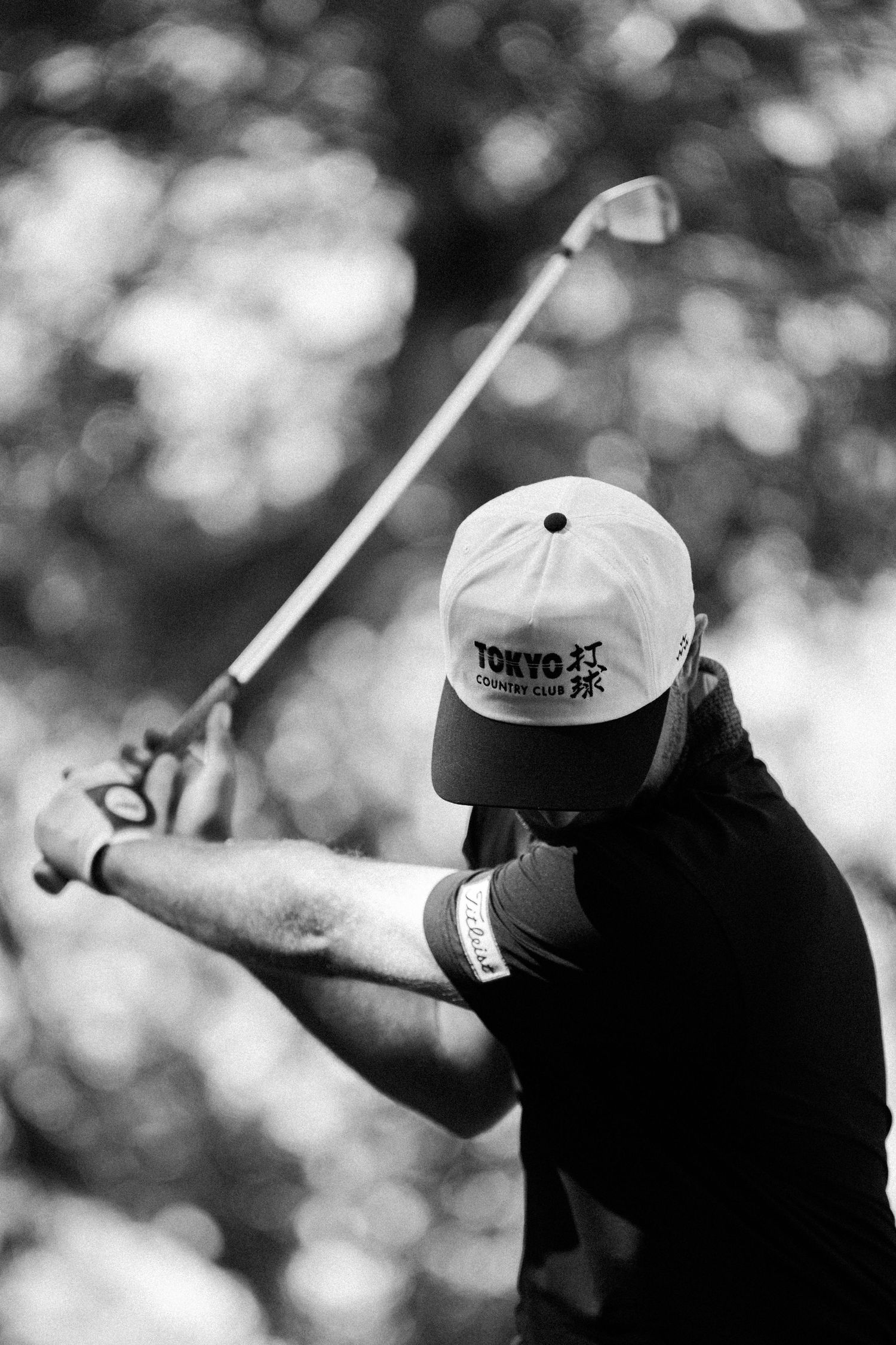 Black and white photo of a golfer mid-swing wearing a Tokyo Golf Club cap outdoors.