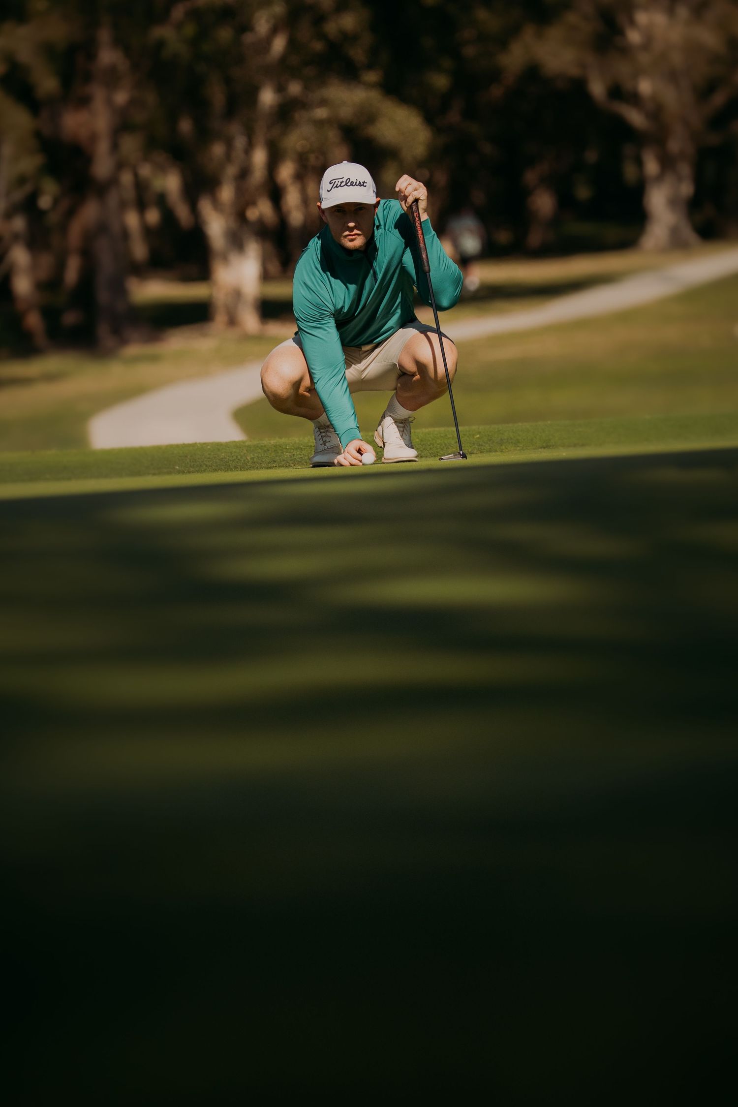 Golfer in teal shirt crouches to line up putt on manicured green golf course.