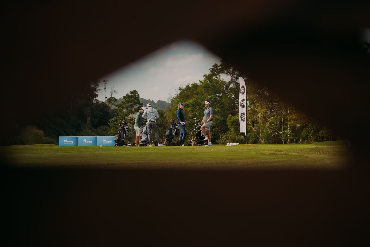 Golfers carrying bags across a lush green fairway framed by trees at a professional golf tournament.