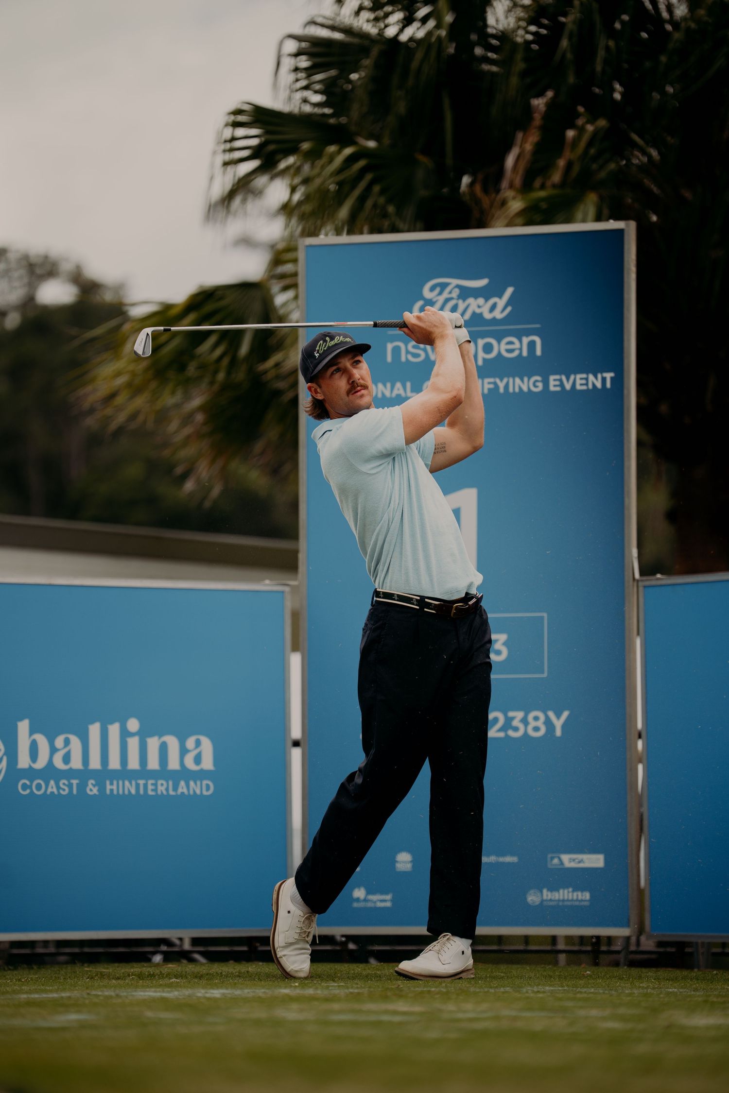 Golfer mid-swing at Ford NSW Open final qualifying event, Ballina Coast and Hinterland backdrop, wearing white shirt and dark pants.