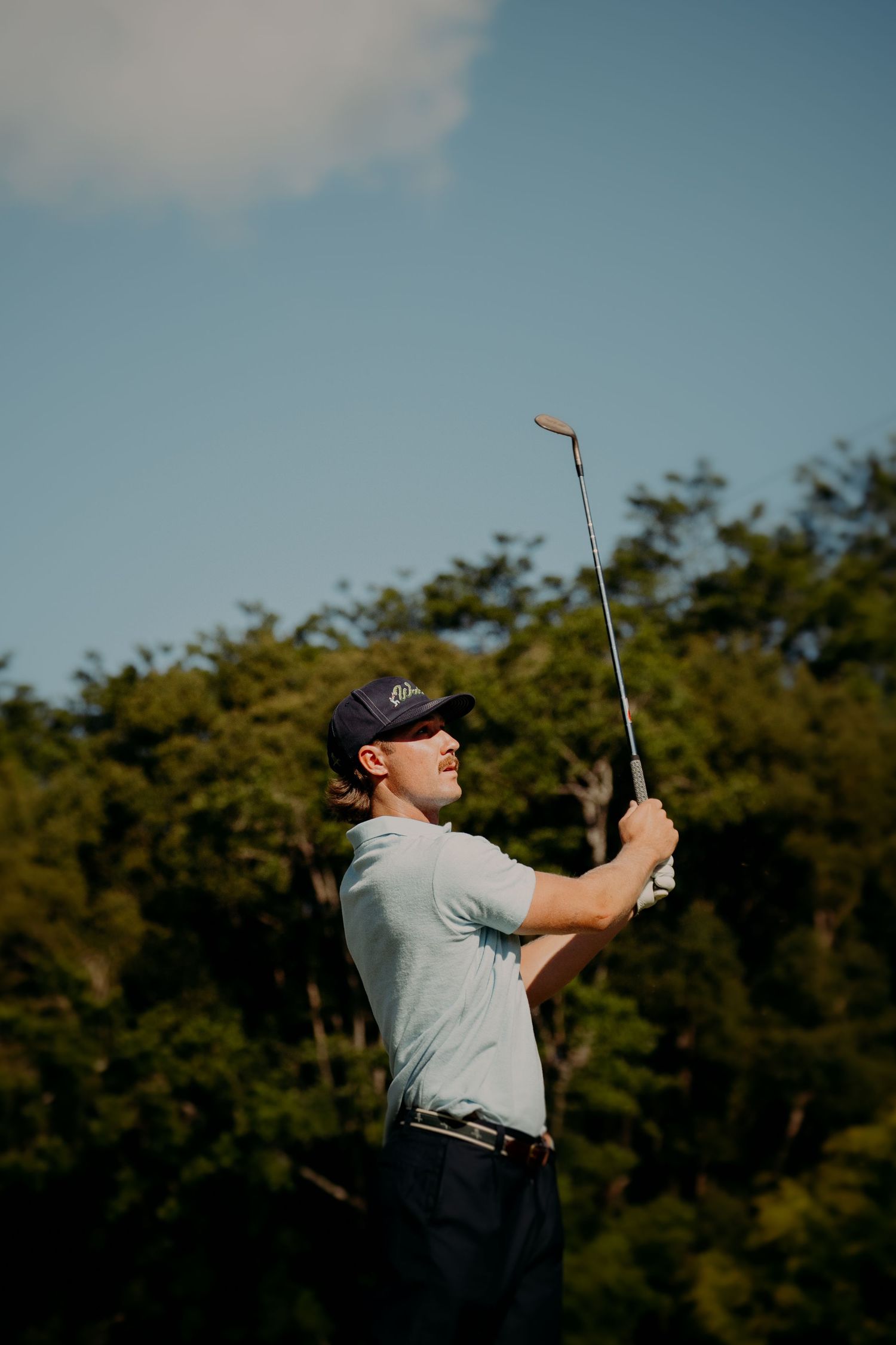 Golfer in blue cap and polo shirt holds club aloft after swing on sunny golf course.