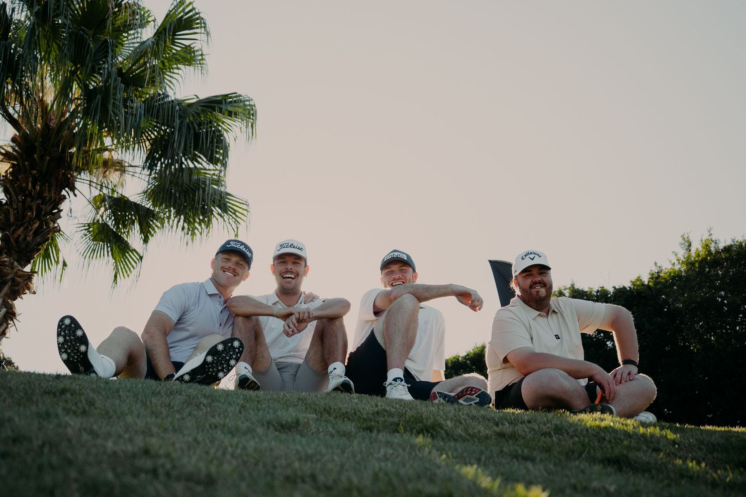 Four men in white shirts and caps sitting on a grassy hill near a palm tree outdoors.