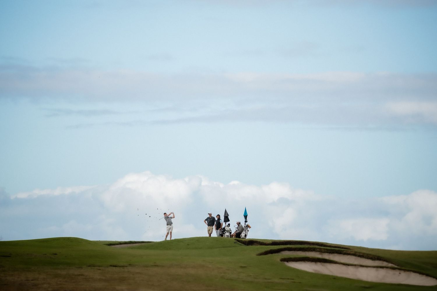 Golfer swinging on a lush green golf course with sand bunkers, caddies, and a blue sky with clouds in the background.