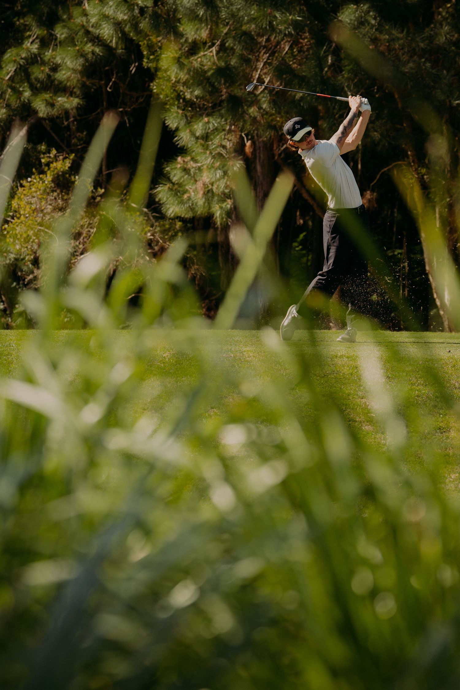 Golfer completing a swing on a lush green golf course surrounded by trees, viewed through foreground grass.