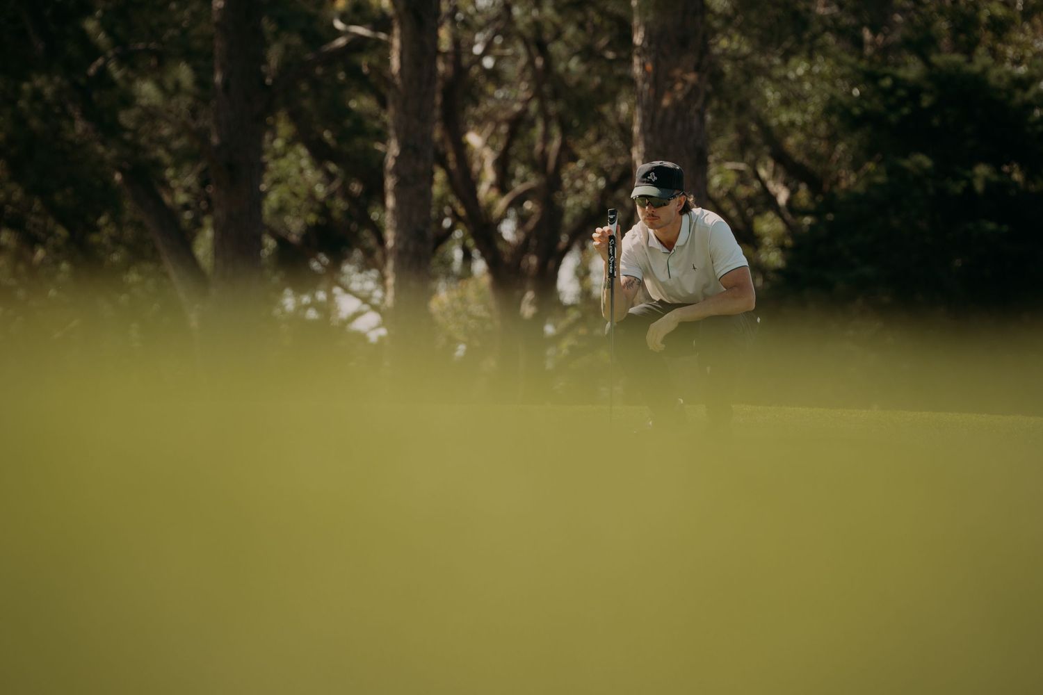 Golfer crouching on a lush green course, carefully reading the putt with trees in the background.