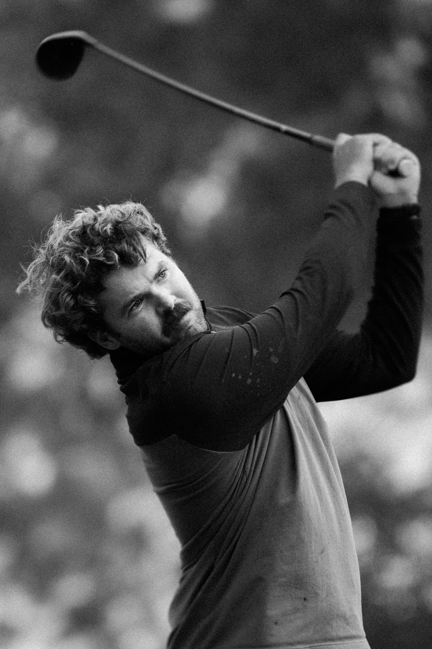 Black and white photo of a curly-haired man mid-swing with a golf club during a tournament.
