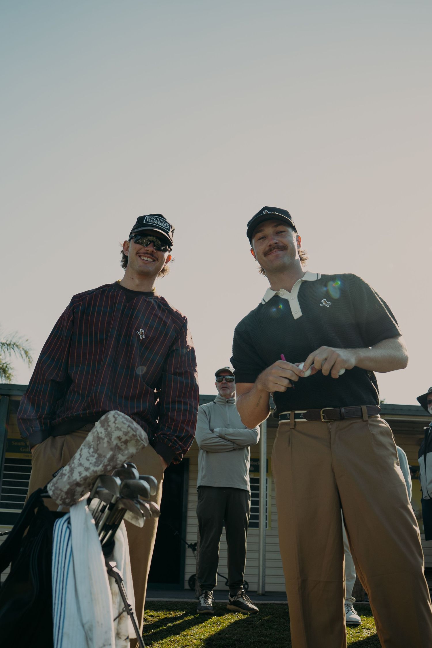 Two men in vintage golf attire and caps smile on a golf course, one holding clubs, with a third person visible in background.