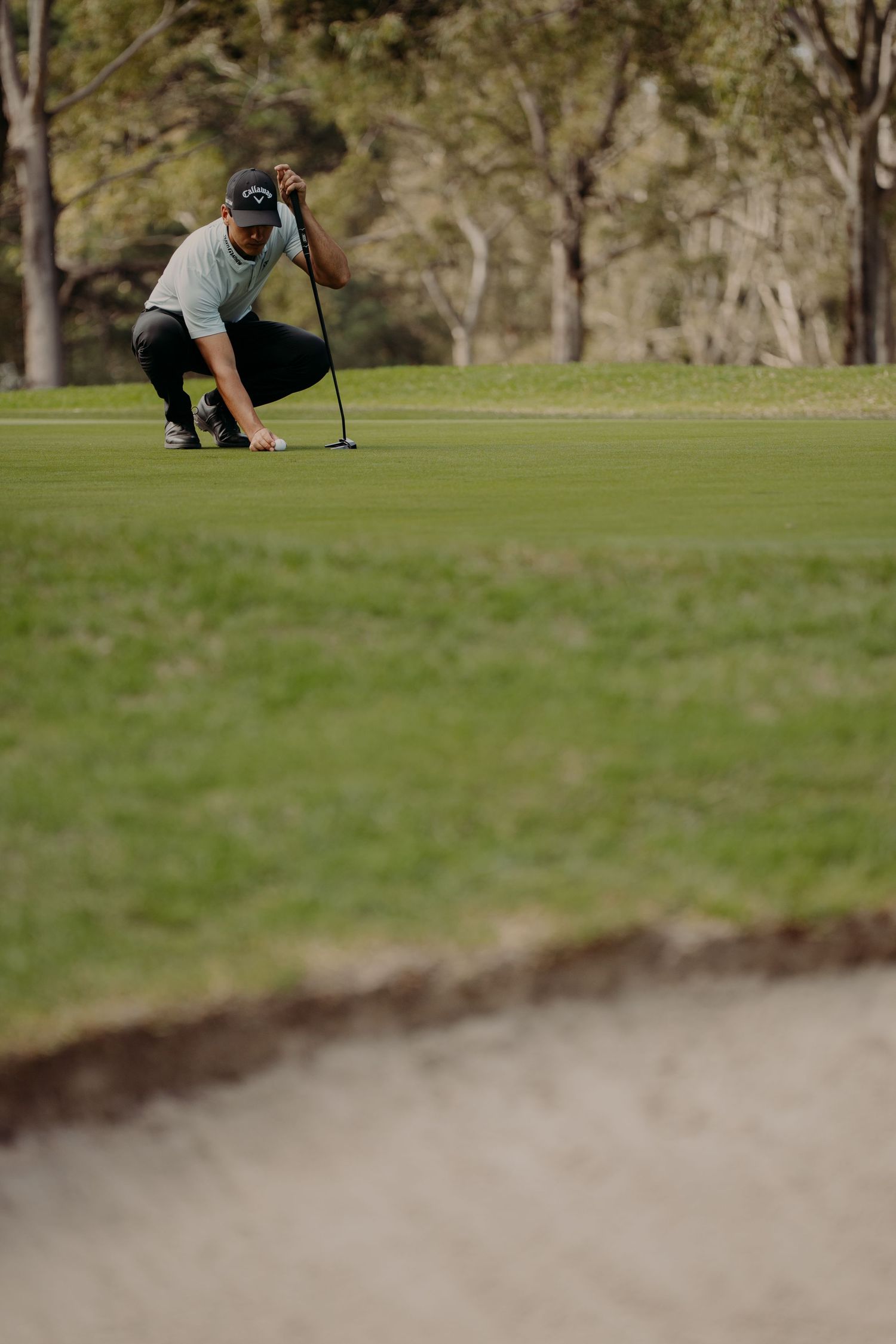Golfer crouching on a green to read the putt, holding a club on a sunny golf course.