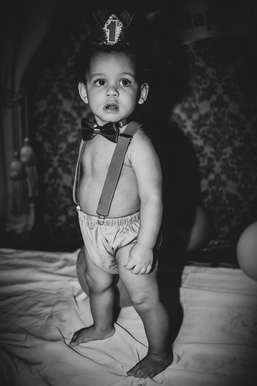 Black and white photo of a toddler boy wearing suspenders, bow tie, and a crown celebrating his first birthday.