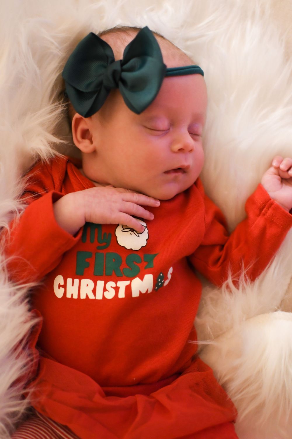 Sleeping newborn baby in red My First Christmas outfit with green bow headband on fluffy white blanket.