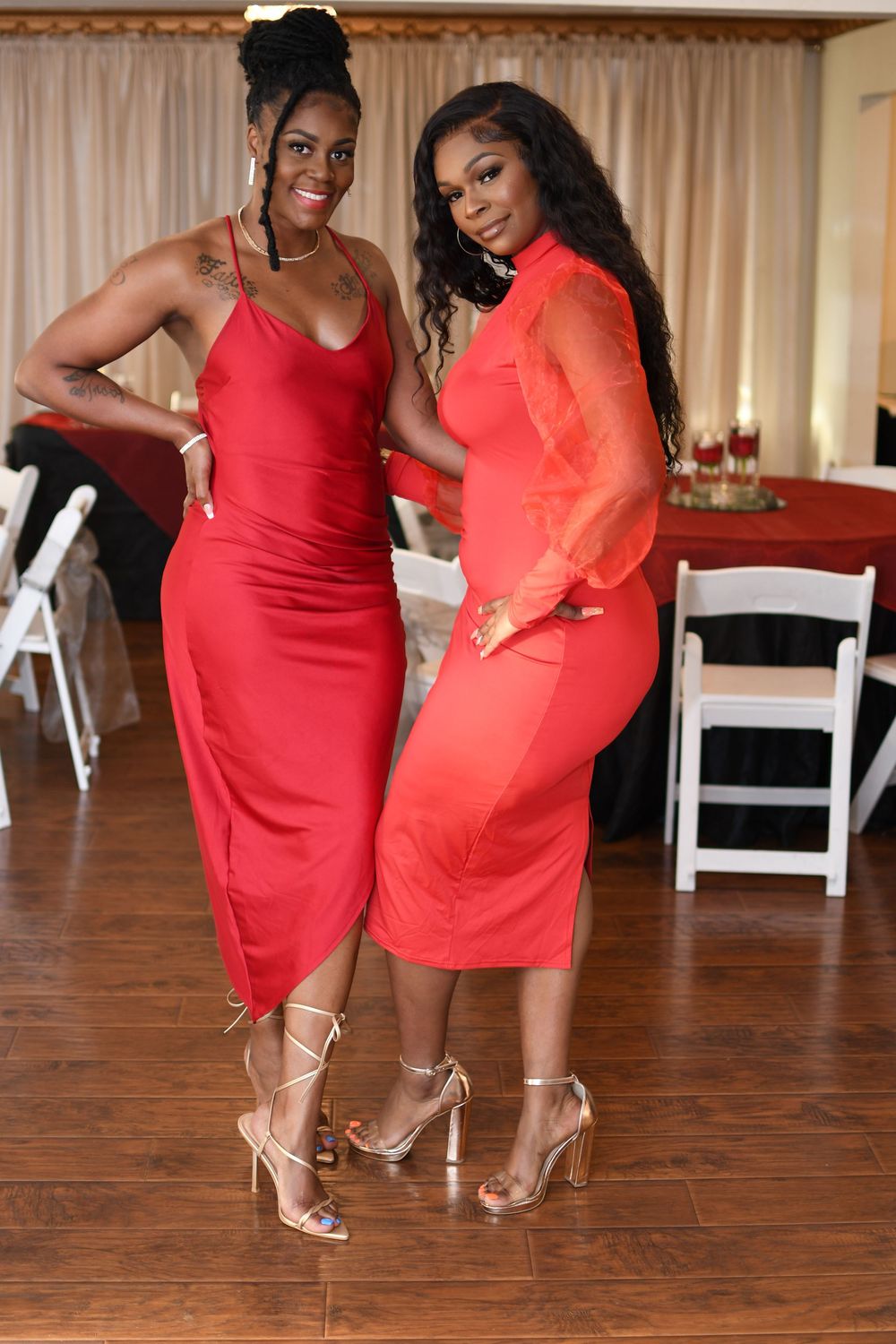 Two women in red dresses and heels pose together at an elegant event venue with white chairs.