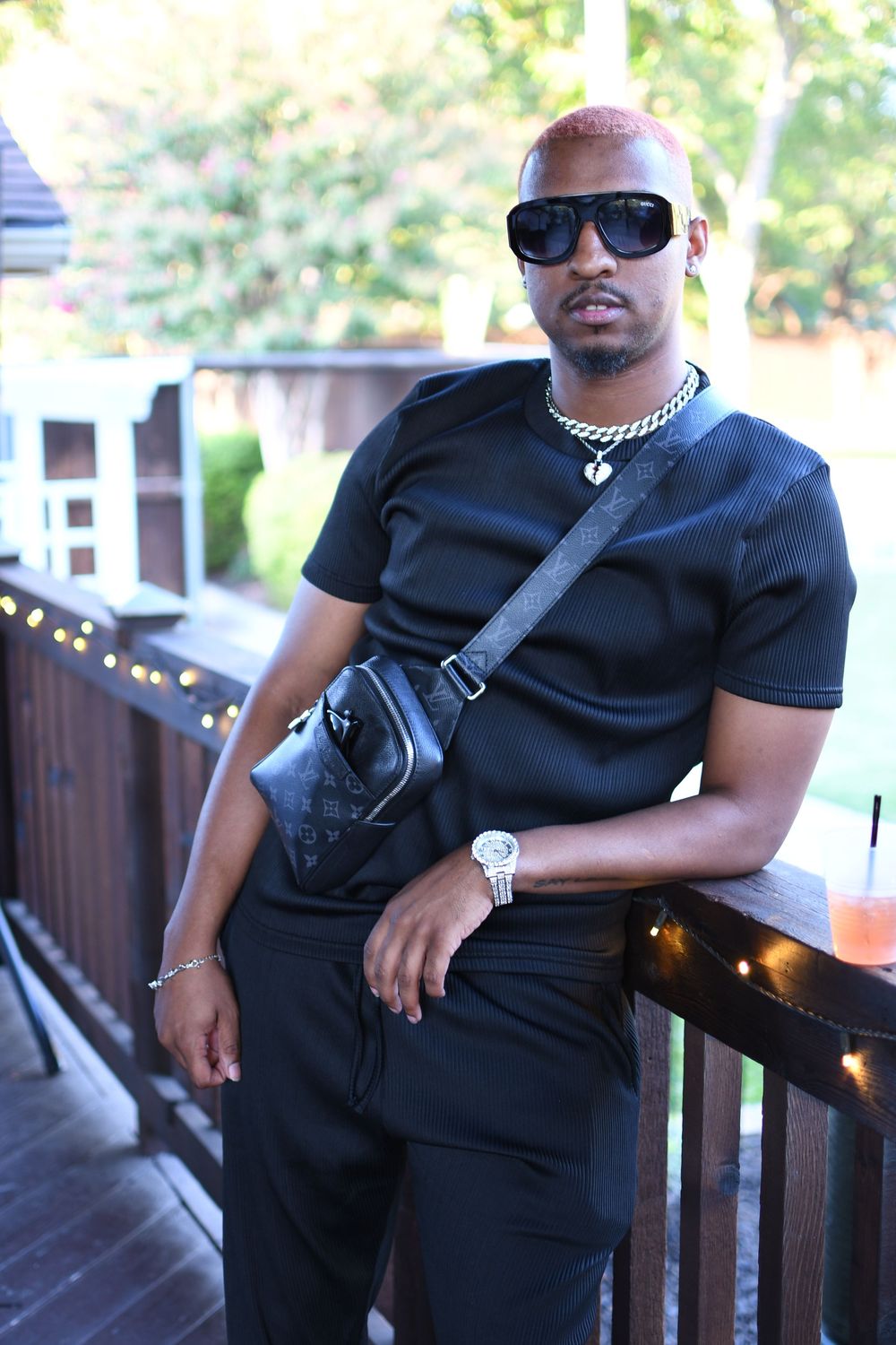 Stylish man in all-black outfit with designer crossbody bag, gold chain, watch, and sunglasses leaning on wooden railing.