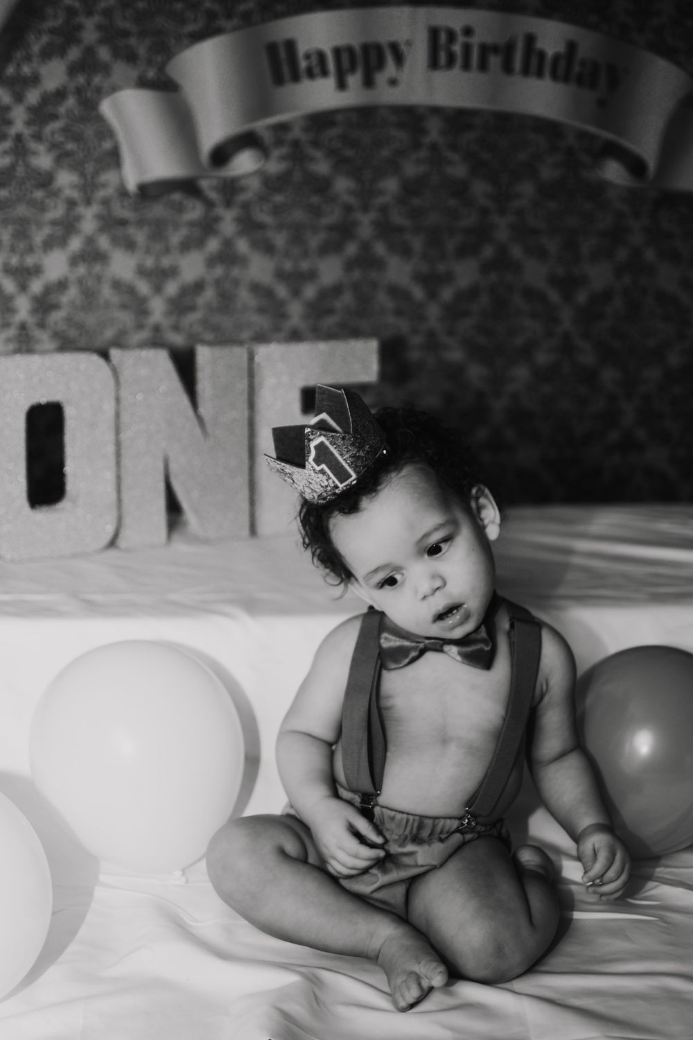 Black and white photo of a baby in suspenders and bow tie sitting with balloons celebrating their first birthday.