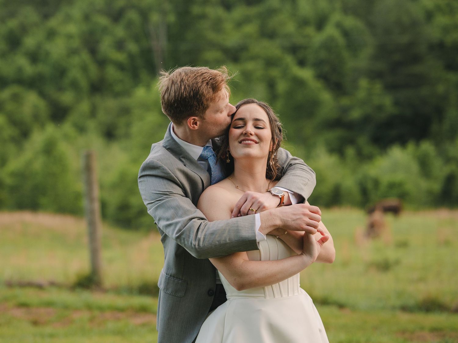Groom in gray suit kissing bride on cheek outdoors in green field, wedding couple embracing joyfully.