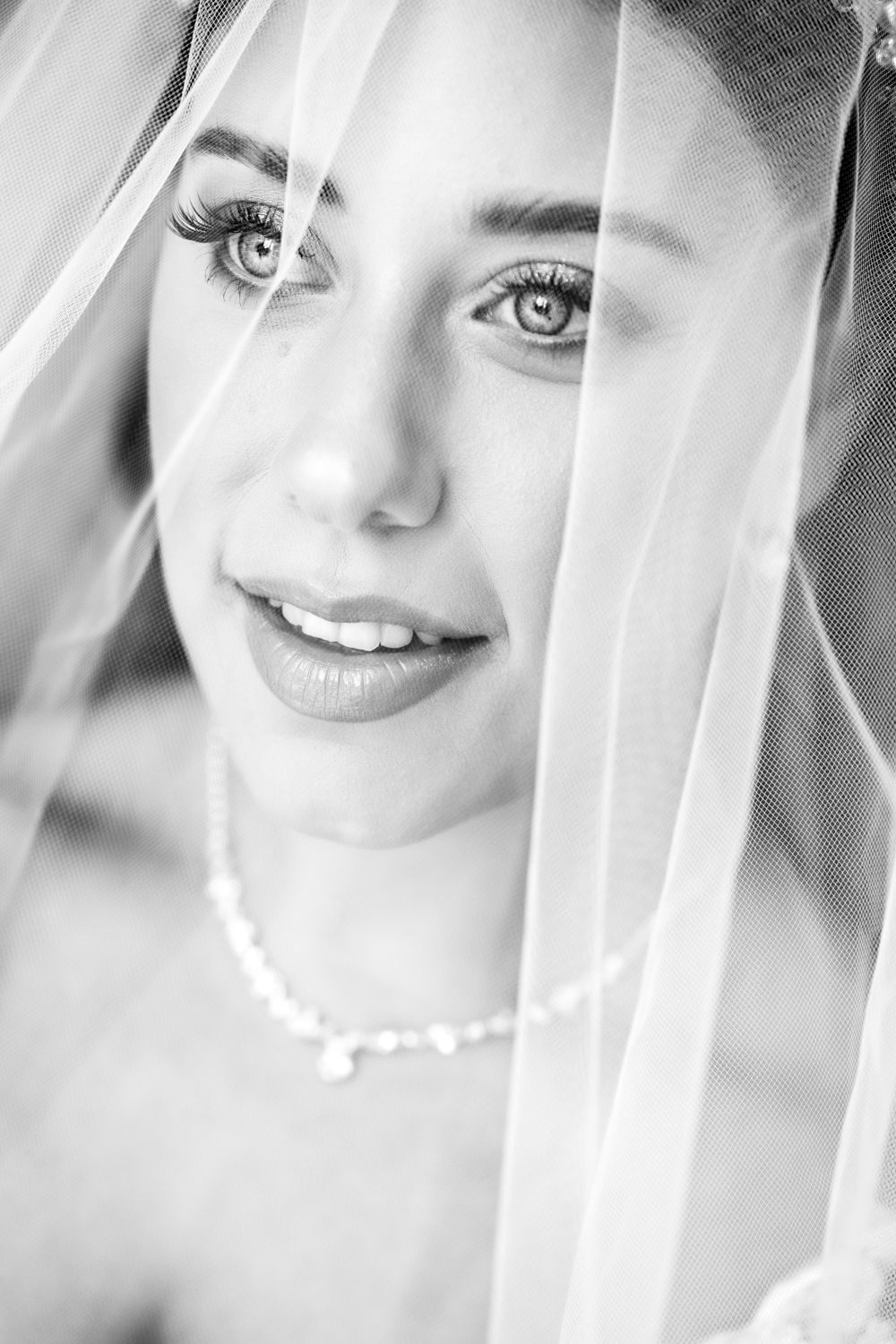 Black and white portrait of a smiling bride wearing a pearl necklace and veil, with beautiful eyes and elegant makeup.